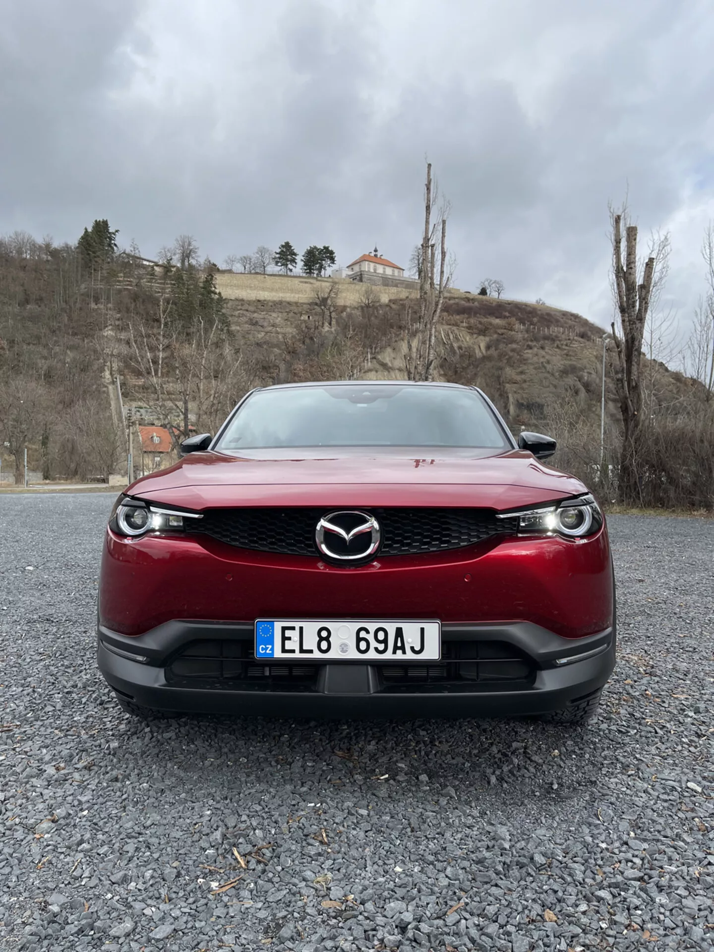 Framifrån vy av en röd Mazda MX-30 från 2020, framhävande bilens grill, stötfångare och LED-strålkastare på en grusyta mot en bakgrund av molnig himmel med grönska och en byggnad