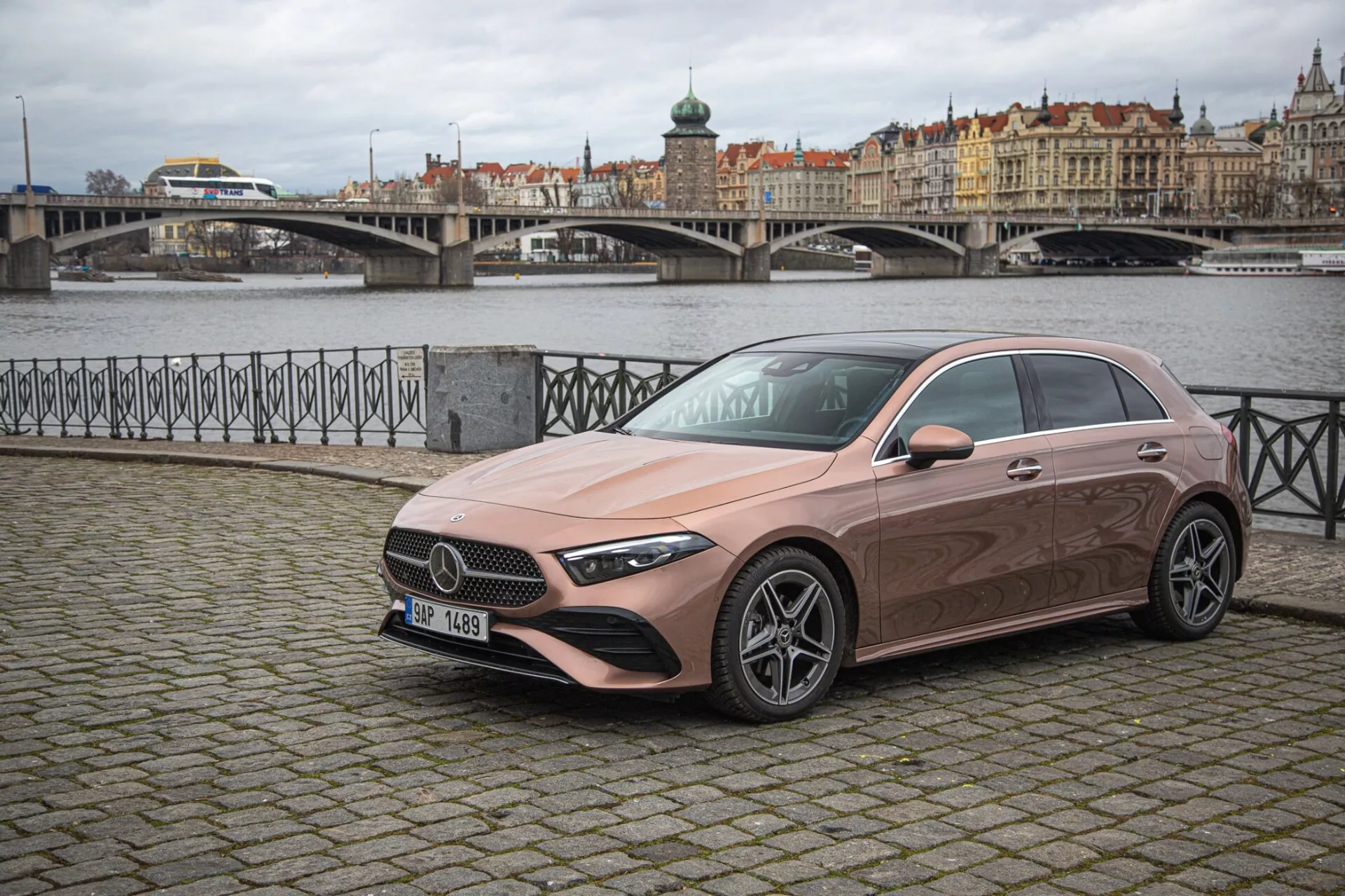 2022 års Mercedes-Benz A-klass fyradörrars sedan parkerad på en europeisk kullerstensgata med en pittoresk stadsvy och vatten i bakgrunden, som visar sin slanka, aerodynamiska design, framträdande grill, LED-strålkastare och metallisk lackfinish.