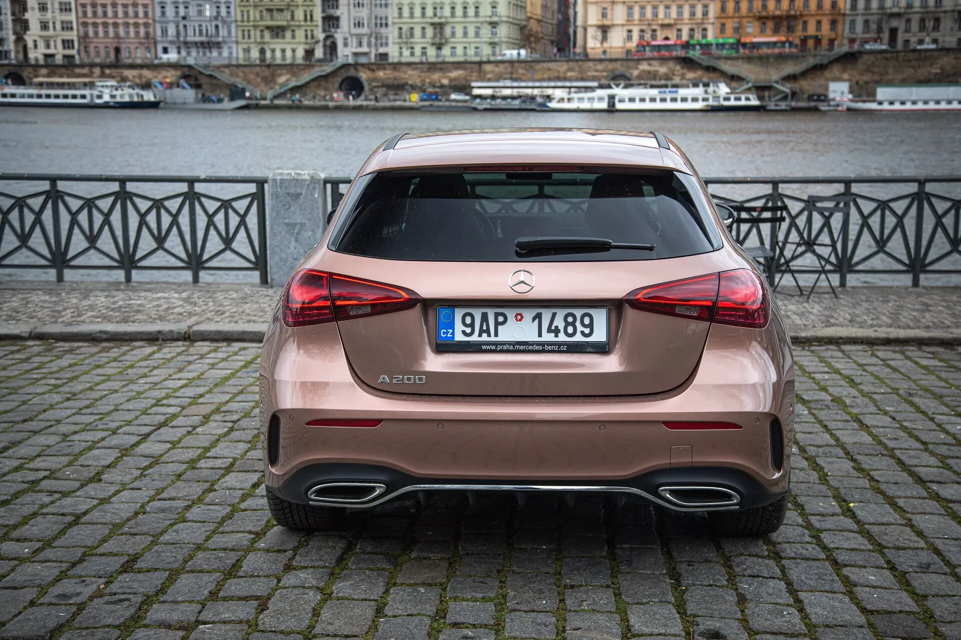 Baksida av en Mercedes-Benz A-klass A 200 parkerad på en kullerstensyta i en stads miljö nära en flod, som visar bilens dubbla avgassystem, halvkombi design och parkeringssensorer