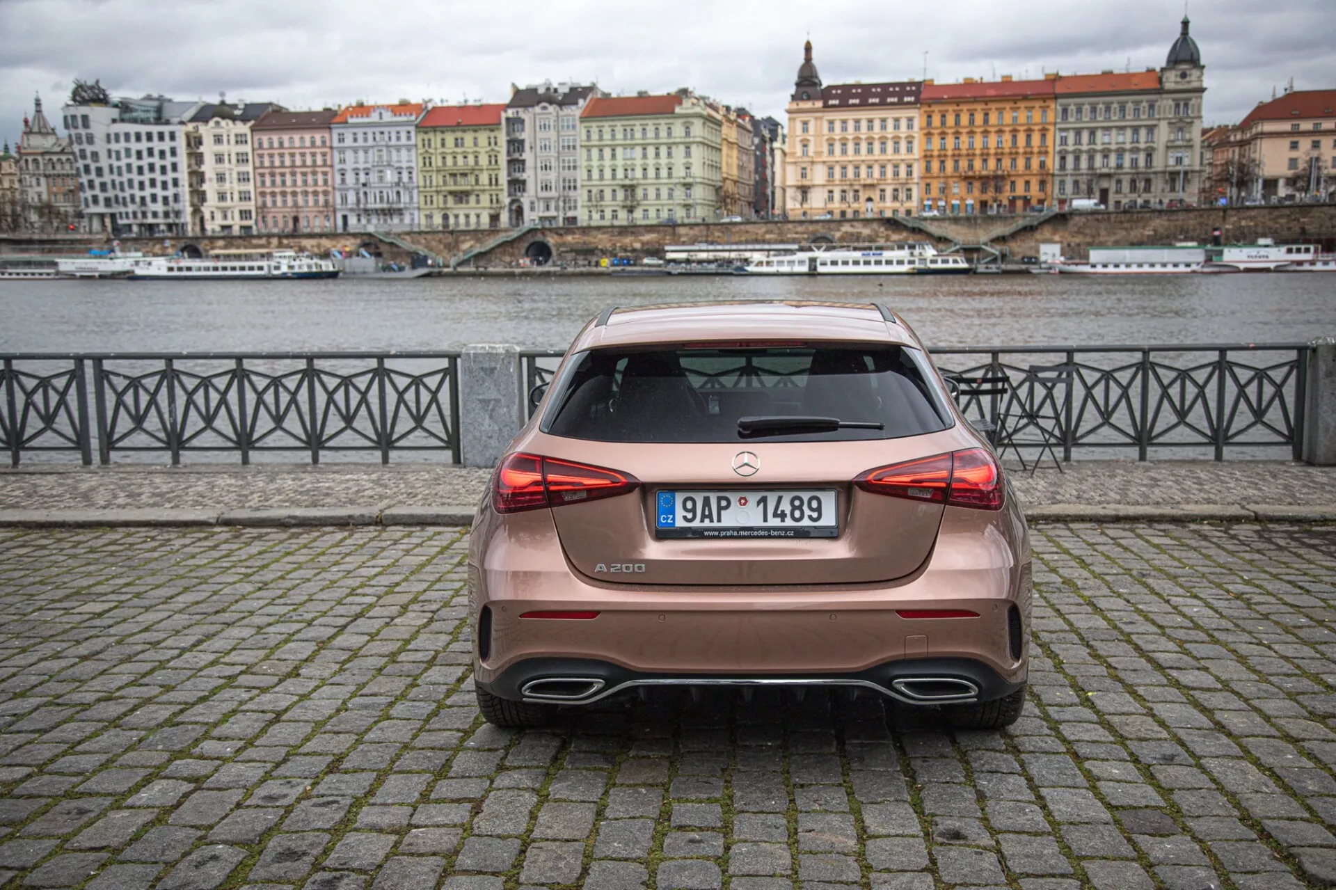 Bakvy av 2022 års Mercedes-Benz A-Klass hatchback parkerad på kullerstensbelagd yta med vatten och byggnader i bakgrunden under en molnig himmel, med LED-bakljus, en takspoiler och dubbla avgasrör