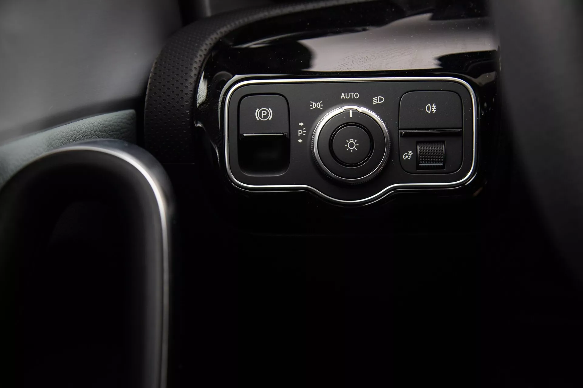Närbild av strålkastarkontrollpanelen på Mercedes-Benz A-Klass 2022 på instrumentbrädan, med knappar för parkeringsljus, automatiskt system, och dimljus eller strålkastarnivelleringsknappar, samt en kontrollratt för ljusinställningar, innesluten i en blank svart ram.