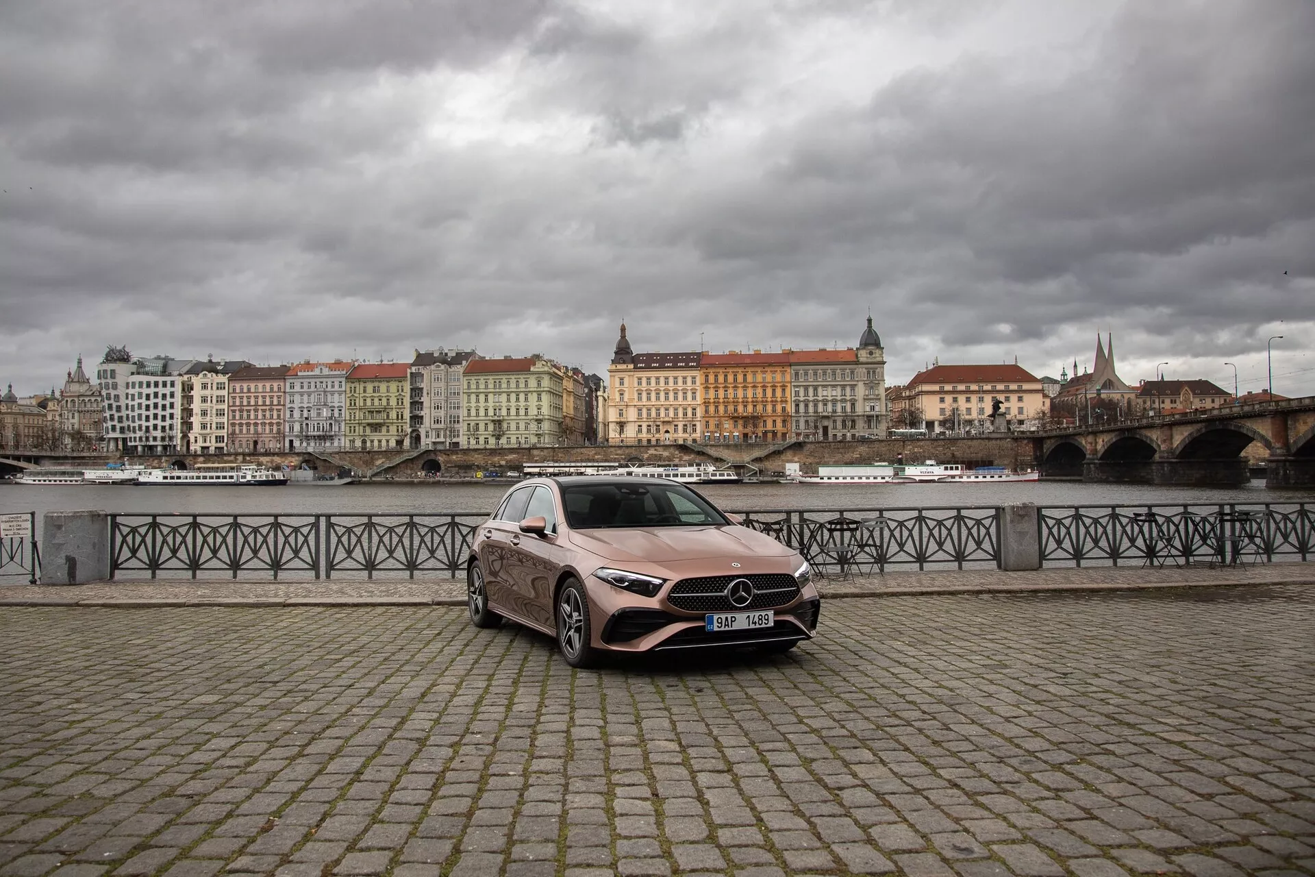 Mercedes-Benz A-Klass från 2022 parkerad på en strandpromenad med kullerstensyta, visar dess främre vänstra kvartal med distinkt grill, LED-strålkastare och stora legeringshjul mot en europeisk stadsbakgrund