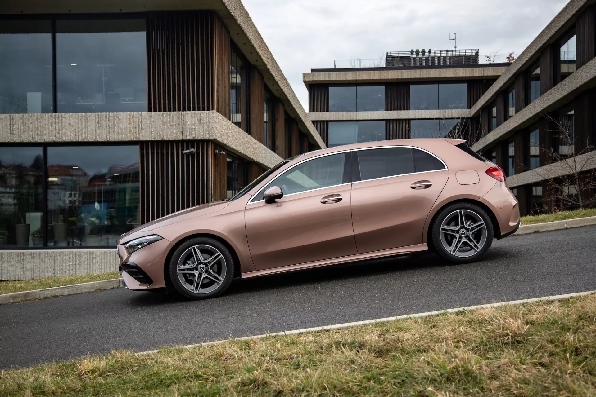 2022 Mercedes-Benz A-Klass sedan i metalliskt brons, parkerad parallellt med trottoarkanten, visar sidoprofil med sluttande taklinje och fälgar med flera ekrar, mot en urban bakgrund med en samtida byggnad
