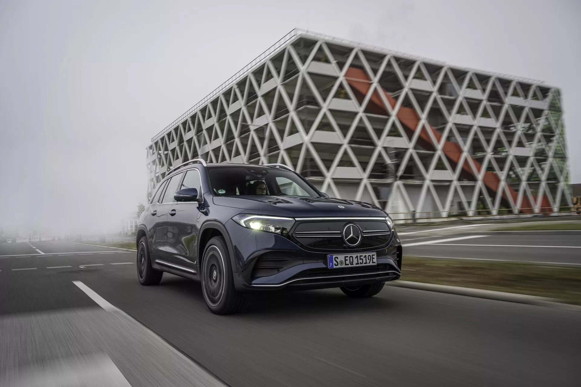 Bilden visar en modern, fyrdörrars Mercedes EQB 2021 SUV i rörelse, fångad från en låg och framsidig vinkel. Fordonet är placerat till vänster och visar sin front, motorhuv och sidoprofil upp till bakdörren. Bilens framhjul är lätt vridet mot betraktaren, vilket antyder en dynamisk manöver. Synliga funktioner inkluderar frontgrillen med en framträdande emblem, LED-strålkastare, dagljuslampor och aerodynamisk design. Den metalliska lackytan reflekterar ljuset och framhäver dess polerade yta. Lättmetallfälgarna ökar estetiken och antyder prestandafunktioner. Bakgrunden inkluderar en futuristisk byggnad med en geometrisk fasad, som framhäver en urban och teknikdriven miljö som överensstämmer med fordonets avancerade tekniska funktioner.