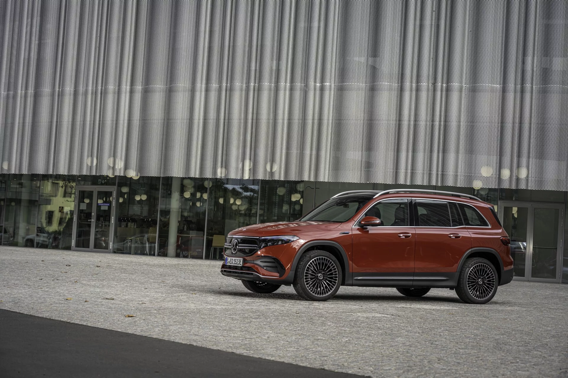 Sidoprofil av en röd SUV, sannolikt Mercedes EQB 2021, fångad från en sida, som visar hela dess längd och designelement som dörrar, fönster, speglar, hjul och kaross. Fordonet står på en asfalterad yta med en modern byggnad med reflekterande glasfasad och metallridåvägg i bakgrunden, vilket antyder en urban miljö. Bilden är tagen i naturligt dagsljus med mjuka skuggor, vilket tyder på en mulen himmel eller skugga från direkt solljus. SUV-en har en elegant design, sannolikt utrustad med avancerade navigationssystem, ADAS, infotainmentsystem, bränsleeffektivitet eller alternativ drivning och lyxiga detaljer som panoramasoltak och lädersäten.
