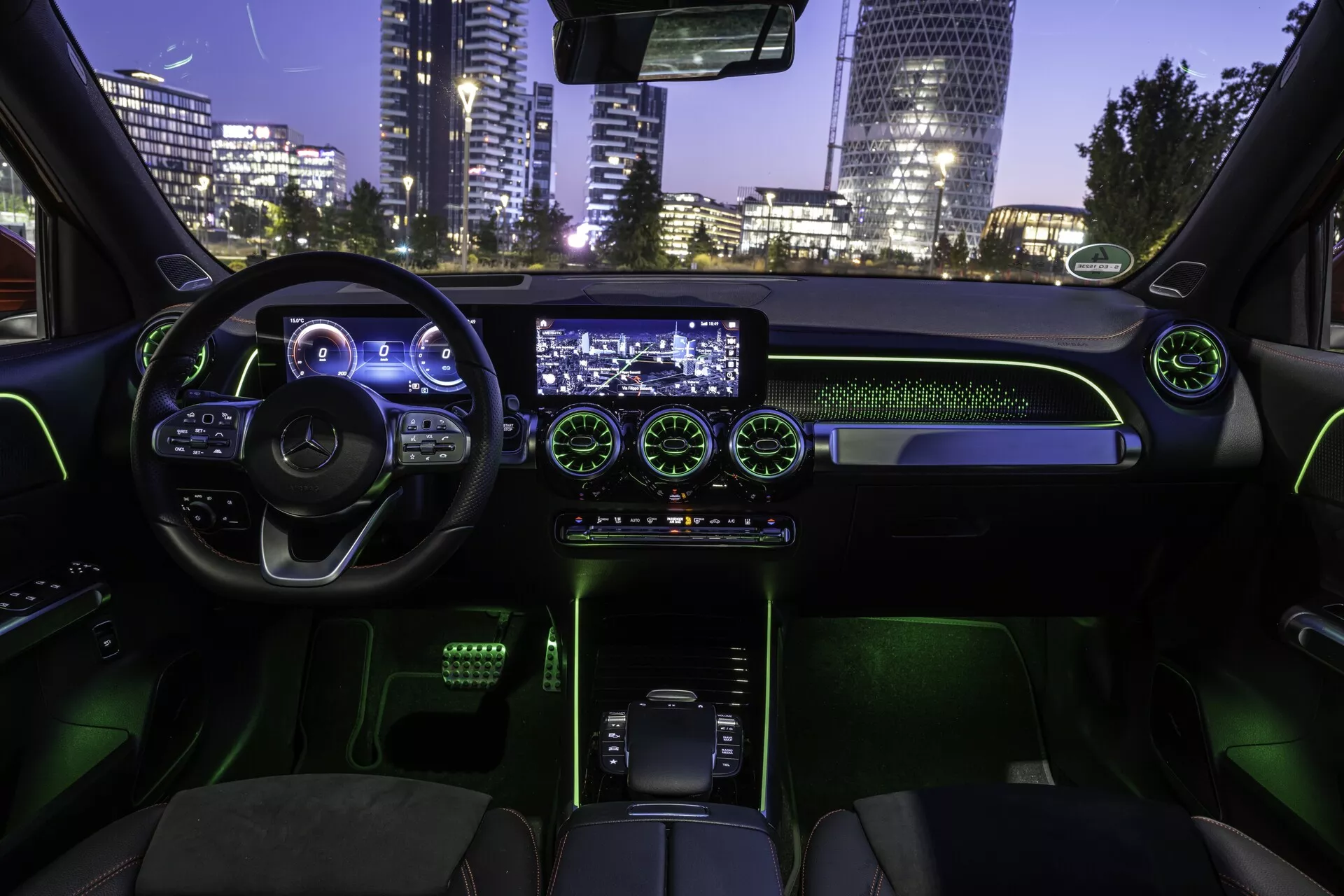 Interiörbild av 2021 Mercedes EQB från baksätet, med vy framåt mot främre kabinen. Den ergonomiska ratten med flera kontrollknappar, platt botten och upplyst digital instrumentpanel skapar en högteknologisk atmosfär. Den centrala infotainment-skärmen och de belysta runda luftventilerna bidrar till en modern känsla. Nedre konsolen har en modern växelspak omgiven av grön bakgrundsbelysning. Klädda säten och grön ambient belysning skapar en futuristisk känsla. Stadslandskapet som syns genom vindrutan antyder en nattlig urban miljö.