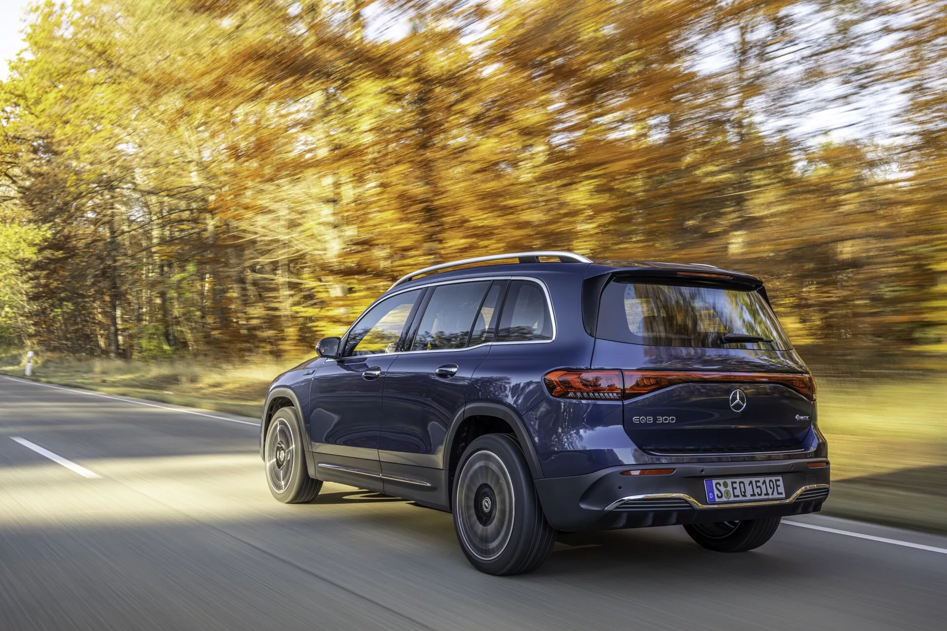 Dynamisk vy av en 2021 Mercedes EQB i rörelse på en trädkantad väg. Den bakre trekvartsvinkeln visar höger sida av el-SUV:en med modern och elegant design, metallisk finish och upphöjd hållning. Höjdpunkter inkluderar bakljus, bakre stötfångare, lättmetallfälgar, sidodörrar och sluttande taklinje. Rörelseoskärpa antyder hastighet och den ljusa dagmiljön bidrar till den exklusiva estetiken.