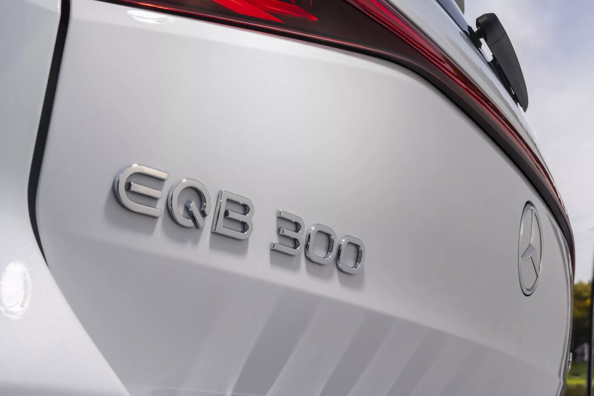 Närbild i diagonal vinkel av bakdelen på en 2021 Mercedes EQB 300, som framhäver emblem och logotyp. Bilden visar den treuddiga stjärnan i en cirkel, modellbeteckningen 'EQB 300', en del av bakluckan och den vänstra baklyktan. Kameravinkeln är från en lägre synvinkel och riktad uppåt, vilket lyfter fram märkningen, moderna designelement, LED-teknologi och den premiummässiga byggkvaliteten hos detta helt elektriska fordon. Bilens kaross är i en glansig vit färg, vilket framhäver dess lyxiga utseende.