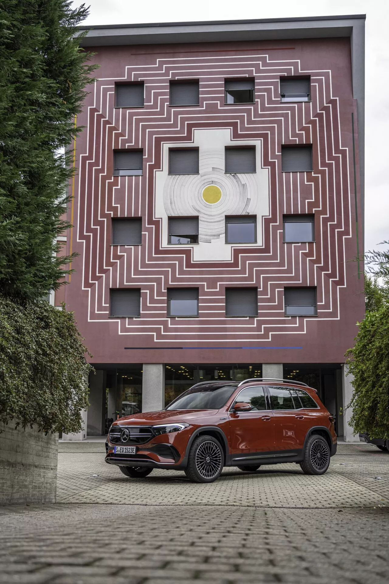 Bild av en modern SUV, sannolikt en lyxmodell, parkerad på ett område med kullerstensliknande textur i en urban miljö eller gårdssetting. Bilen är vänd mot kameran i en vinkel som visar dess framträdande grill med ett centralt emblem, LED-strålkastare, aerodynamiskt designade sidobackspeglar, lättmetallfälgar och framstötsfångare med parkeringssensorer. Bakgrunden inkluderar en arkitektoniskt slående byggnad med ett labyrintliknande mönster kring centrerade fönster.