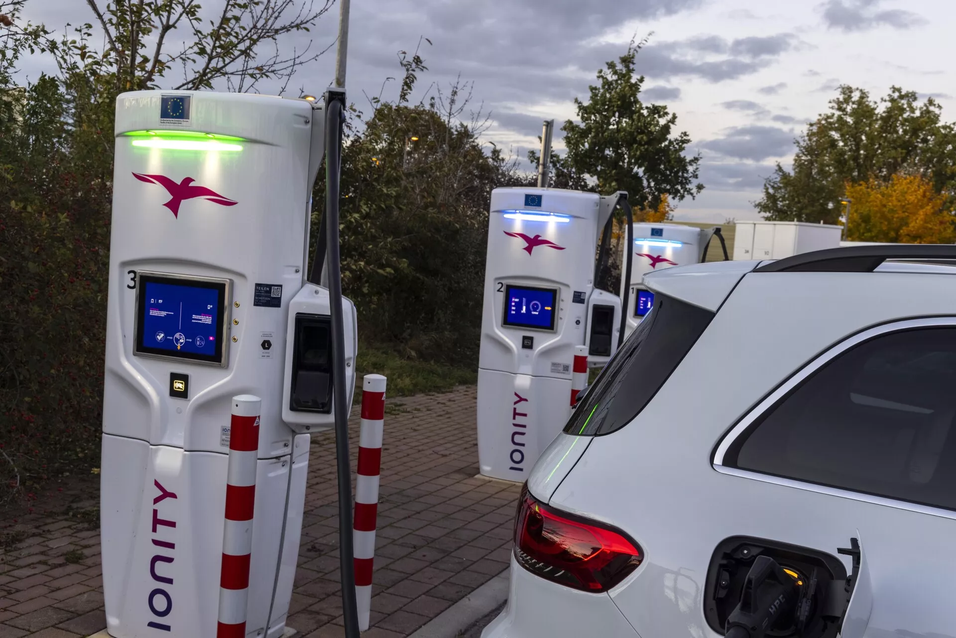 Bilden visar en vit bil, delvis synlig från vänster bak, under en laddningssession vid en laddstation. Fotot, taget från en lätt vinkel mot förarsidan, framhäver den öppna laddporten belägen ovanför bakhjulet och bakom passagerardörren. Det vänstra bakljuset, bakrutan och en del av taket antyder en hatchback eller SUV-kaross. I bakgrunden syns två IONITY-laddningsenheter med blåa skärmar och ett grönt ljus på en, vilket indikerar laddningsaktivitet. Omgivande belysning tyder på kväll eller mulna förhållanden och framhäver bilen som huvudobjektet i ett utpekat laddningsområde.