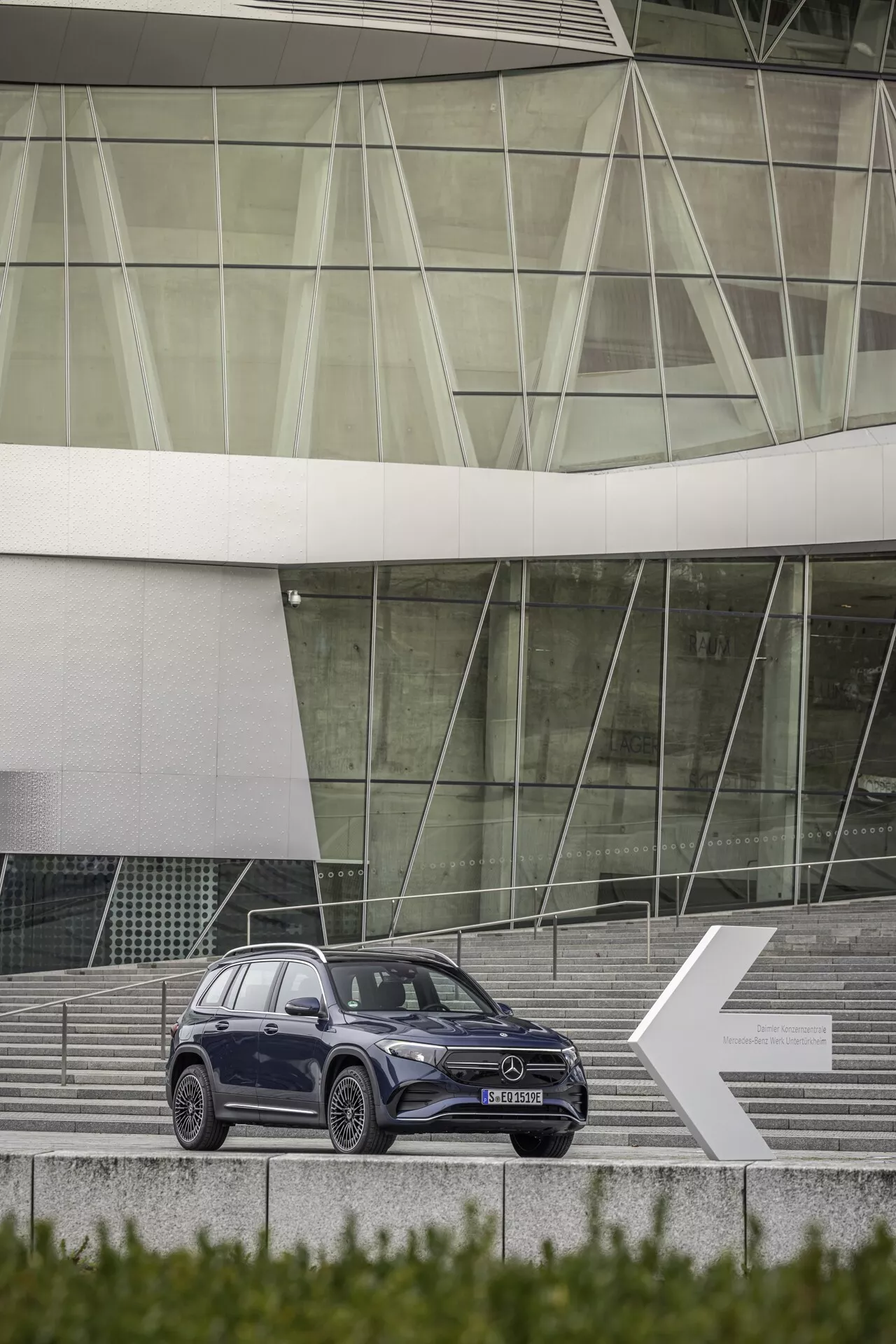 Full sidoprofil av elegant 2021 Mercedes EQB SUV placerad på trappor som leder till en modern byggnad med glasfasad. SUV:n är centrerad och något vinklad åt vänster, visar dess högra sida med designlinjer, flerekrade legeringshjul, sidospeglar, fönster och dörrhandtag. Mörk nyans kontrasterar vackert mot metalliska detaljer.