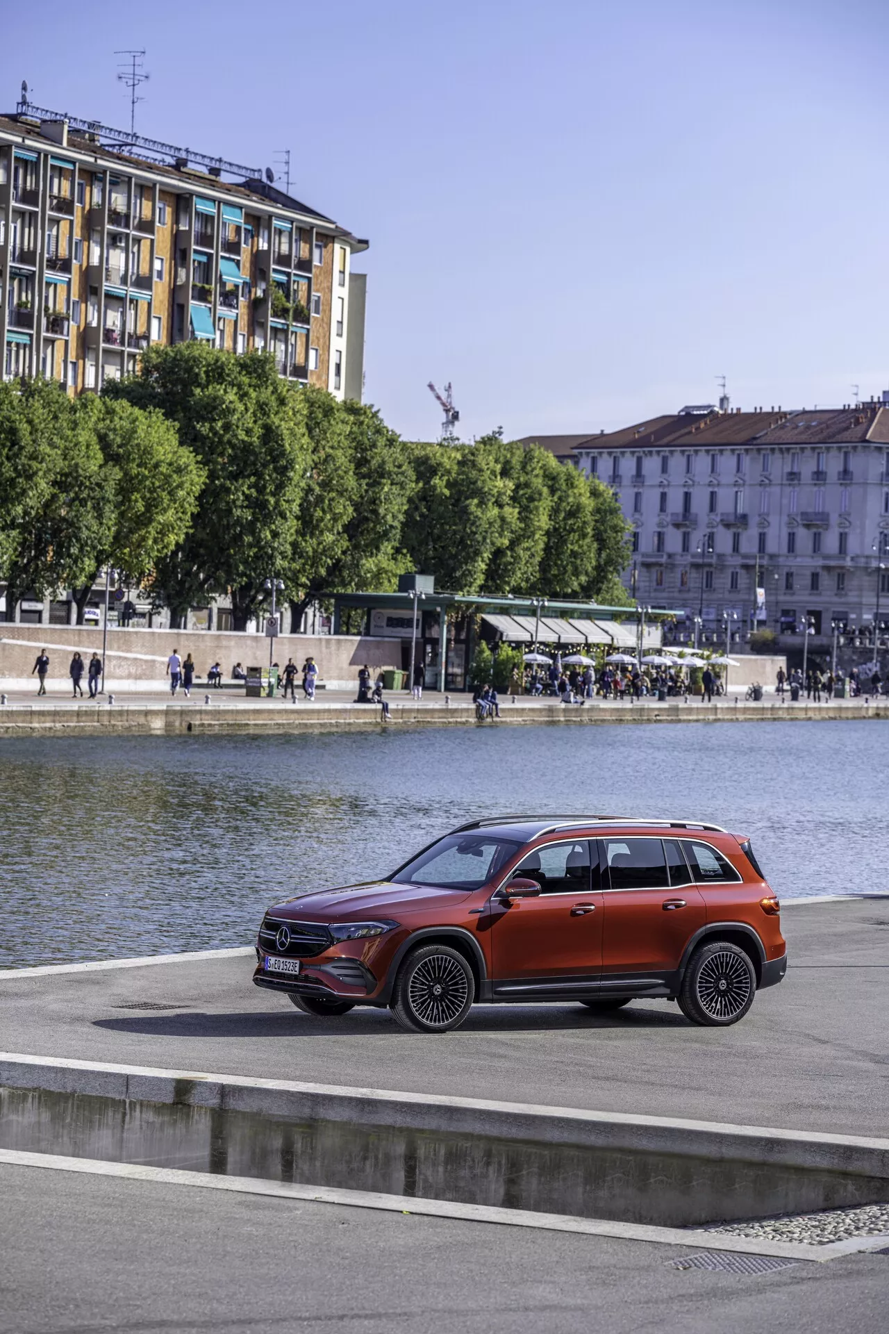 Profilvy av en Mercedes EQB 2021 SUV parkerad vid en vattensida med urbana byggnader och spårvagnshållplats i bakgrunden, som visar upp sin moderna design, eleganta strålkastare, framträdande frontgrill, väl definierade hjulbågar och glansig metallic-finish från en upphöjd vinkel.