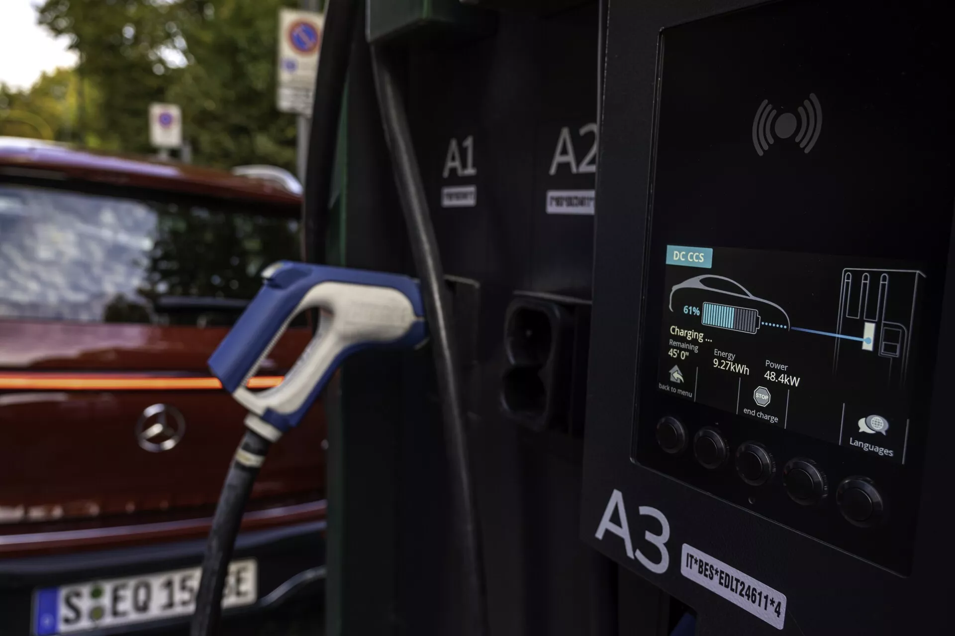 Närbild av en laddstation för elfordon med en bil som laddas i bakgrunden. Laddstationen visar detaljerad information på sin digitala display som laddstatus, energi i kWh och laddningstid. Bilen, en röd eller vinröd halvkombi, är delvis synlig med sin bakre ljusgrupp och baklucka, i en utomhusmiljö med naturligt ljus och lövverk.