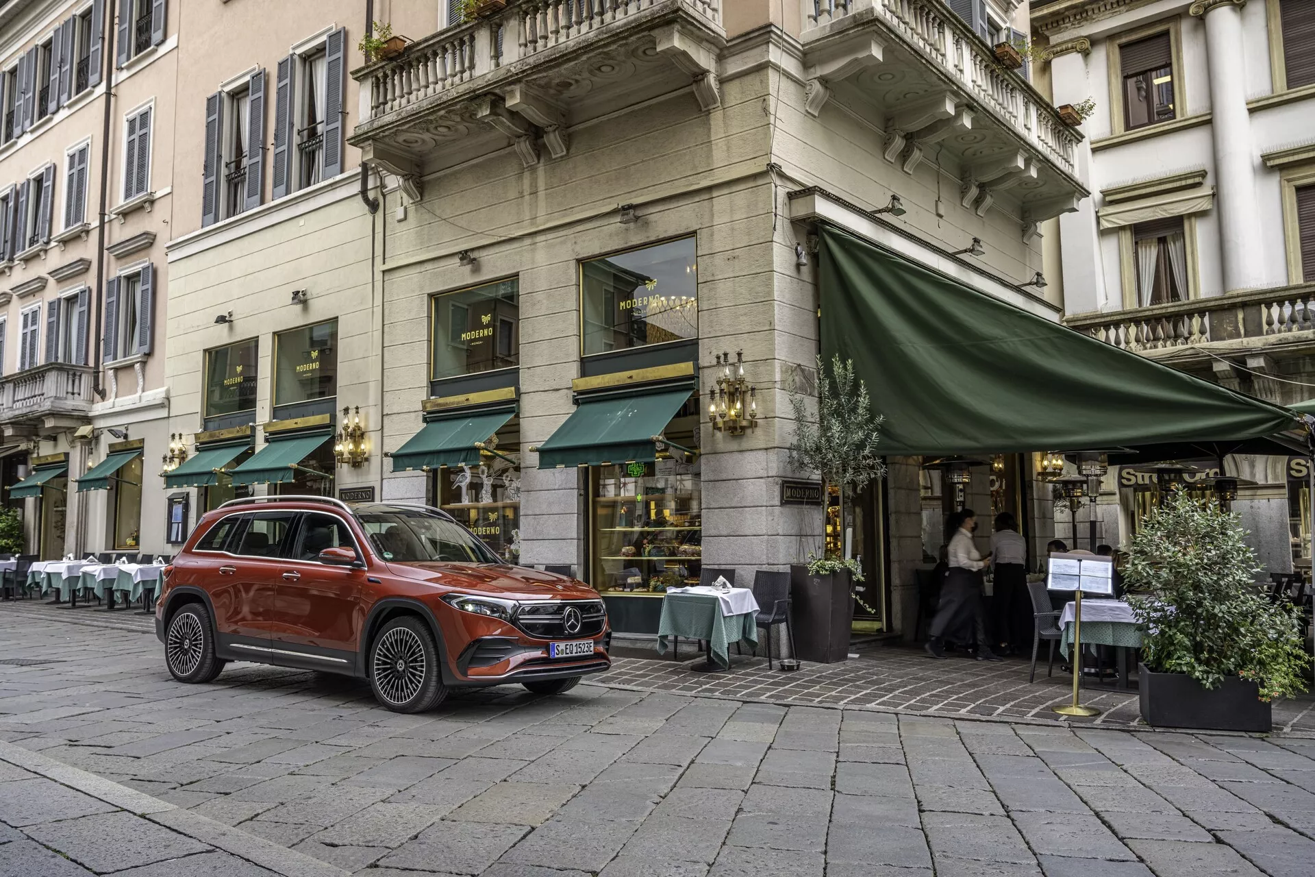 Bilden fångar en fullständig vy av en Mercedes EQB 2021 parkerad på en kullerstensgata framför ett kafé med uteservering under en grön markis. Fotograferad från ett trekvarts främre perspektiv visar den tydligt fordonets främre galler, strålkastare, huva, hjul, främre förardörr och en del av baksidan. Denna moderna kompakta SUV visar designinslag som förhöjd körhöjd, lättmetallfälgar, LED-dagljus och en metallisk lackfinish. Den urbana miljön, förmodligen i en europeisk stad, betonar dess användbarhet som en stadsfamiljebil med potentiella avancerade tekniska funktioner såsom förarassistanssystem.
