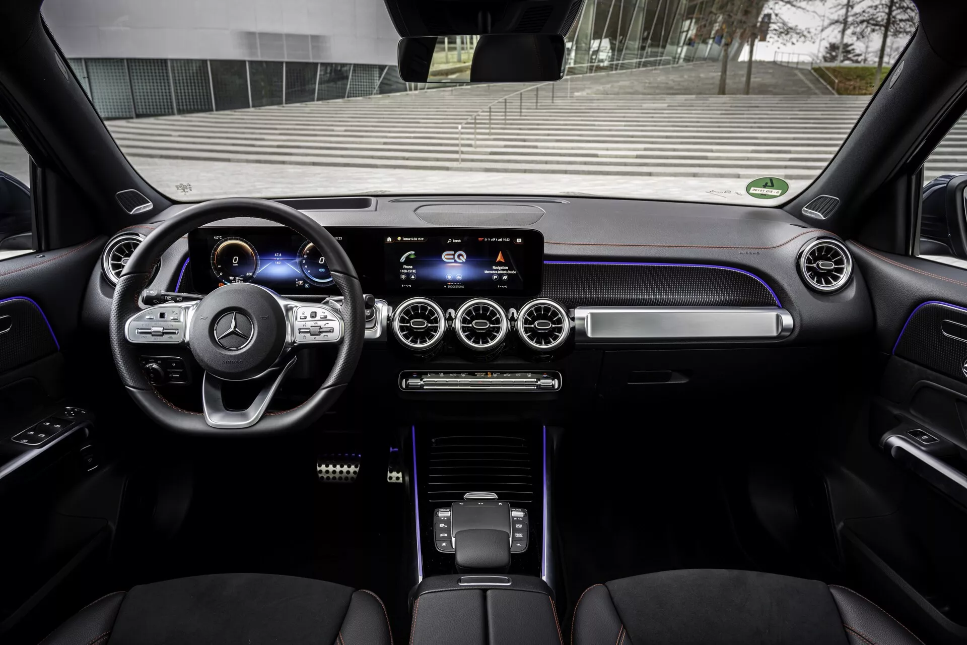Interiör av en modern Mercedes EQB 2021 från förarens perspektiv. Bilden ger en tydlig vy över instrumentpanelen, treekrad ratt med multifunktionsknappar, digitalt instrumentkluster, pekskärms-infotainmentsystem på mittkonsolen, runda luftventiler med metalliska detaljer, taktila klimatkontrollknappar, elegant växelväljare och lyxiga lädersäten. Ambient belysning tillför en premium känsla, med interiören som visar material av hög kvalitet och avancerade tekniska element.