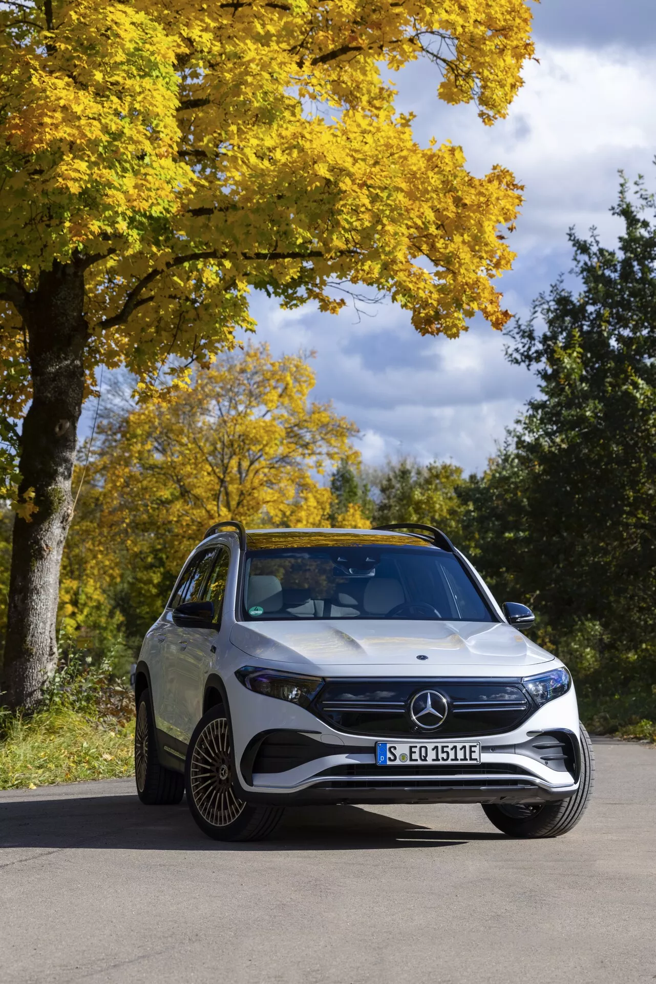 Mercedes EQB 2021 sedd från en vinkel på tre fjärdedelar framifrån, fotad i en naturlig höstmiljö med klar himmel. Den lyxiga el-SUV:en har en framträdande grill med Mercedes-emblemet, LED-strålkastare, sportig främre stötfångare och karaktäristiska linjer på huven. Distinkt flerspokedesign på aluminiumfälgarna och en lutande vindruta syns också, vilket visar på aerodynamisk effektivitet.