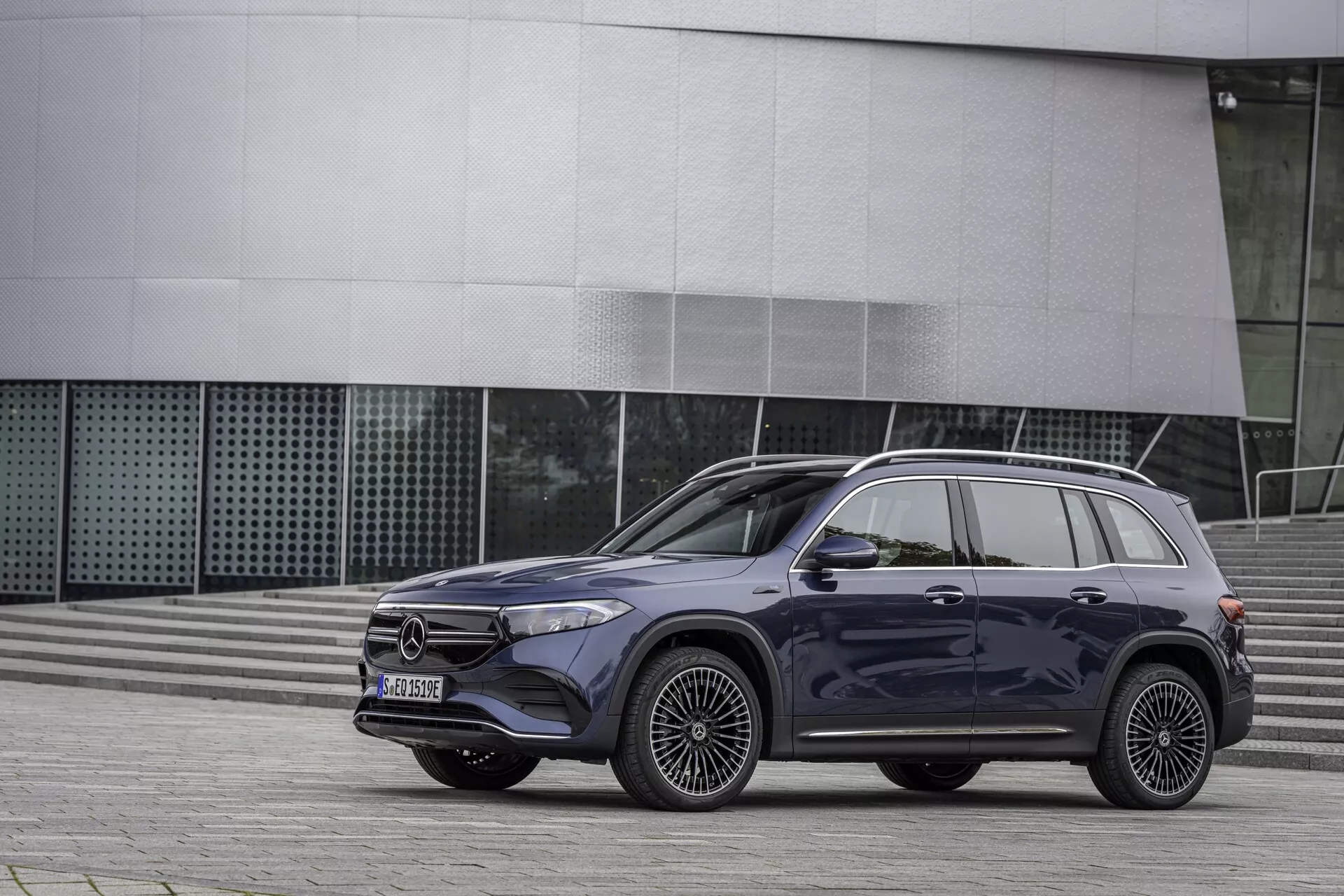 Sidoprofilvy av en Mercedes EQB 2021 SUV parkerad i en urban miljö, som visar en elegant och lyxig design. SUV:n har fyra dörrar med synliga handtag, distinkta legeringshjul, en framträdande frontgrill med ett centralt emblem, LED-strålkastare, kontrasterande sidospeglar, takräcken och ett panoramatak. Sidokjolarna och hjulbågarna förstärker dess robusta utseende. Lågkameravinkeln betonar fordonets dominerande närvaro mot en samtida arkitektonisk bakgrund.