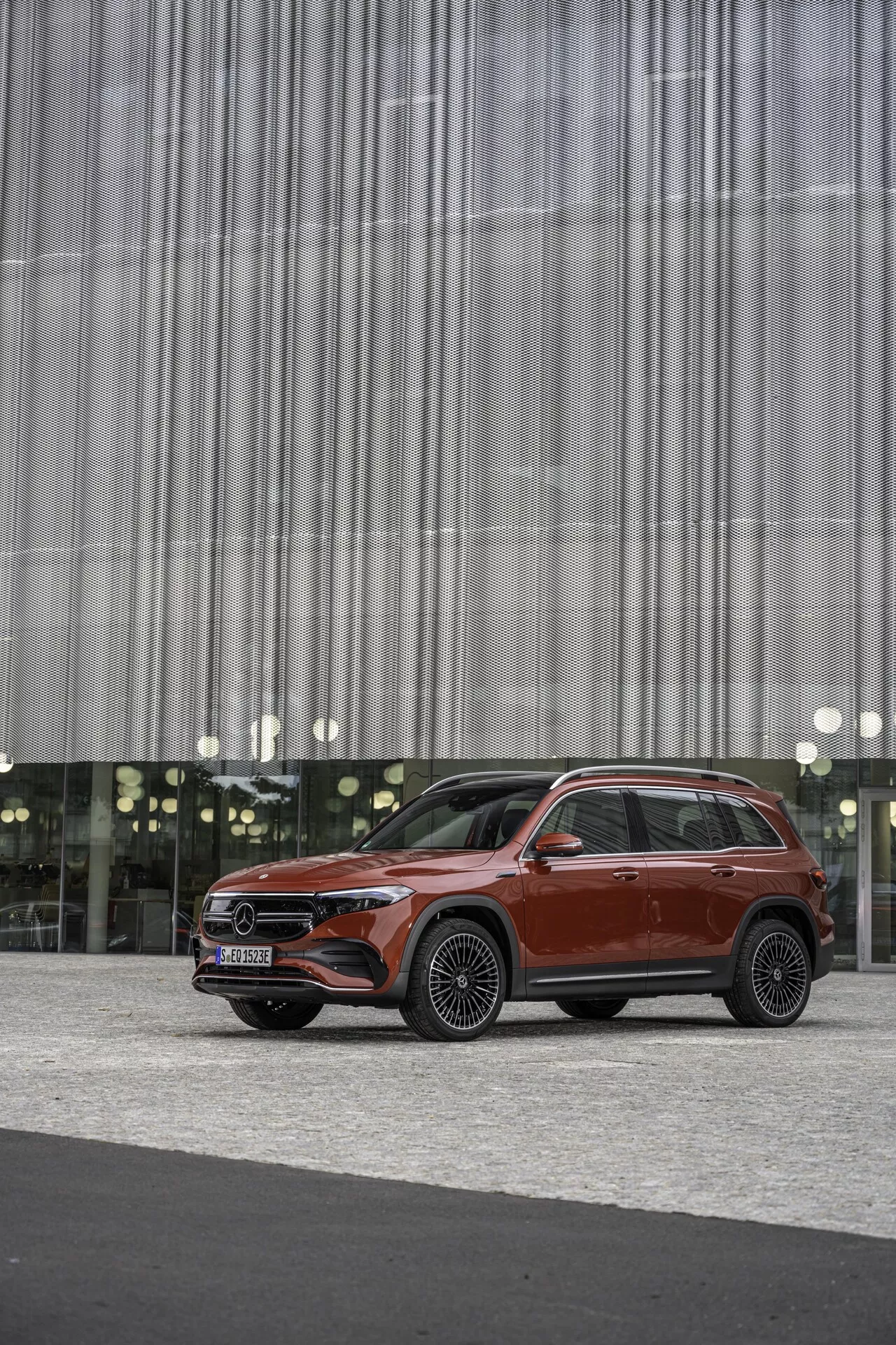 Bild av en modern SUV Mercedes EQB (243) från 2021 i en djup röd nyans, placerad utanför en byggnad med en vertikal linjeformad fasad. Fotograferad från ett trekvartsfrontal-perspektiv, som visar frontgrill med ett framträdande emblem, LED-strålkastare med dagsljusljus, skulpterad motorhuv, stora lättmetallfälgar, hög fönsterlinje, kromade fönsterlister och takräcken. Luksus och nytta blandas i denna samtida bilmodell, framhävd på en neutral bakgrund som betonar designens detaljer.