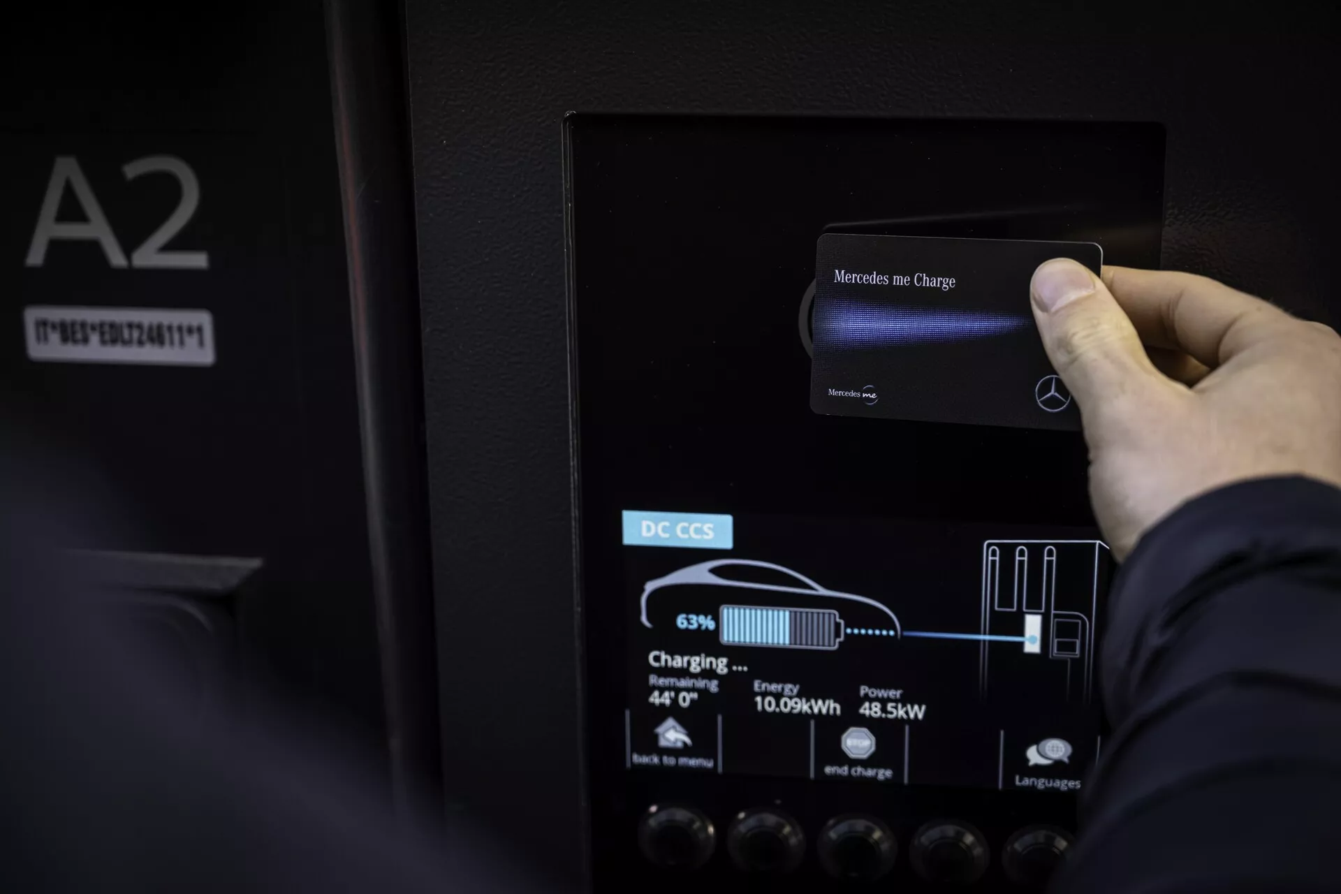 Närbild av en hand som interagerar med pekskärmen på laddstationen för en Mercedes EQB 2021 elbil. Färgskärmens LCD visar 'Mercedes me Charge' högst upp och visar nuvarande laddningsnivå på 63 %, levererad energi på 10,69 kWh och uteffekt på 45,5 kW, med grafiska batteri- och plugindikatorer. Skärmen är monterad på en matt svart panel märkt 'DC CCS', vilket indikerar snabbladdning. Omgivande detaljer inkluderar reflektion av omgivande ljus, panelens textur och en 'A2'-etikett med serienummer till vänster.
