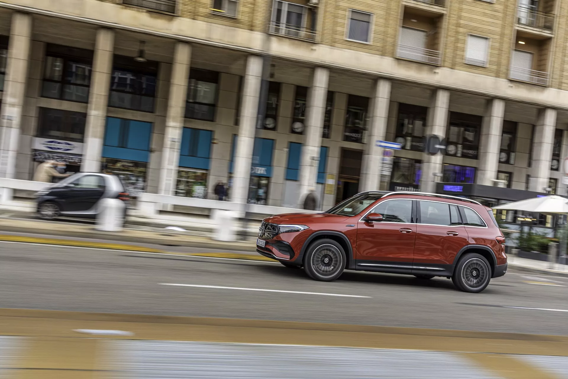 Dynamisk sido sikt av en djupröd eller rödbrun Mercedes EQB 2021 SUV som färdas på en stadsgata. Fångad med panoramafotograferingsteknik, visas SUV:ns profil inklusive dörrar, fönster, hjul, grill och strålkastare med rörelseoskärpa i bakgrunden, vilket framhäver dess hastighet. Bilden lyfter fram fordonets moderna design, lättmetallfälgar och en blank finish i mjukt, överskuggat ljus.