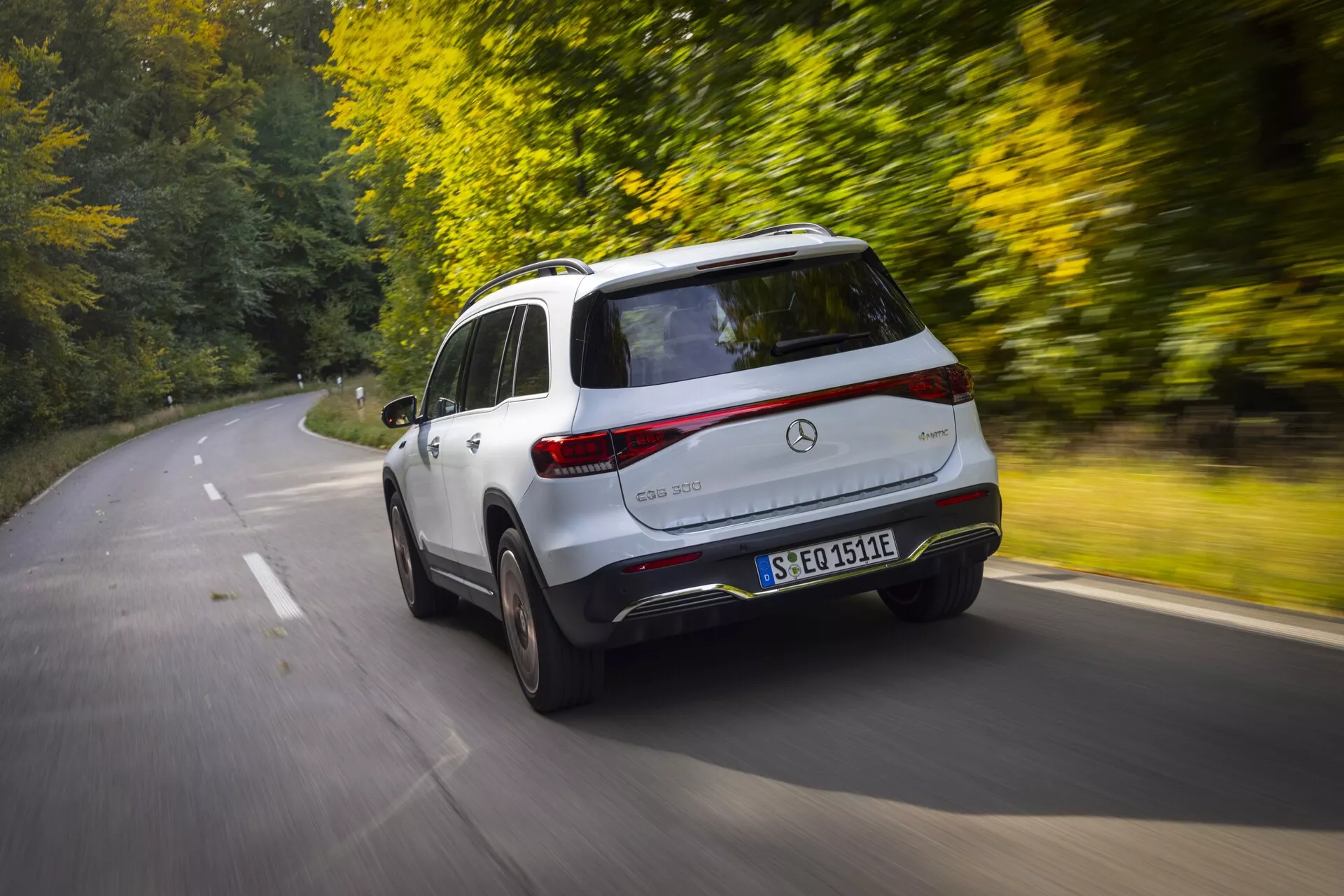 En dynamisk bild av en 2021 Mercedes EQB crossover-SUV i rörelse, tagen från en upphöjd bakre trekvarts vy. Det övervägande vita fordonet visar upp en elegant, aerodynamisk design och betonar de bakre och sidoprofilerna inklusive den vänstra sidan, hela bakre delen, bakrutan, baklyktorna och en del av den bakre stötfångaren. Fordonets distinkta emblem, modellens märkning och registreringsskylten är synliga. Rörelseoskärpa i bakgrunden och på hjulen antyder höghastighetskörning. Den lugna, skogsklädda vägen i höst och solljuset som filtrerar genom träden, ger värme och ger en lyxig, fridfull körupplevelse som är idealisk för resor och äventyr.