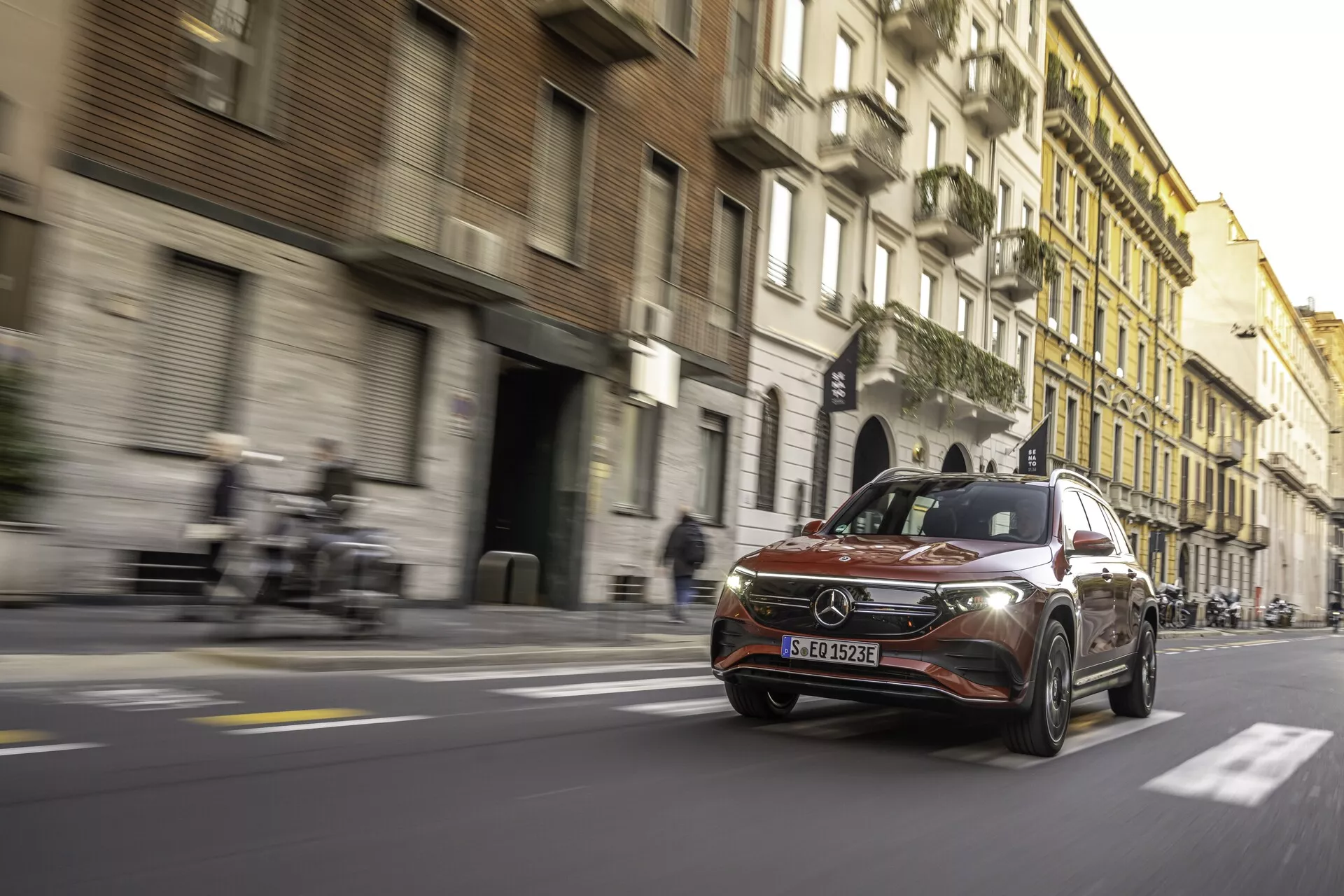 Låg-vinkel foto av en Mercedes-Benz EQB 2021 i rörelse på en urban gata, som fångar främre grillen med emblem, eleganta LED-strålkastare, främre stötfångare, vindruta, del av sidoprofil och moderna aluminiumfälgar. Rörelseoskärpa och ljusstrimmor skapar en dynamisk hastighetseffekt. Bakgrunden inkluderar historiska byggnader som kontrasterar med bilens modernitet. Dagsljusmiljö med varma toner och långa skuggor.