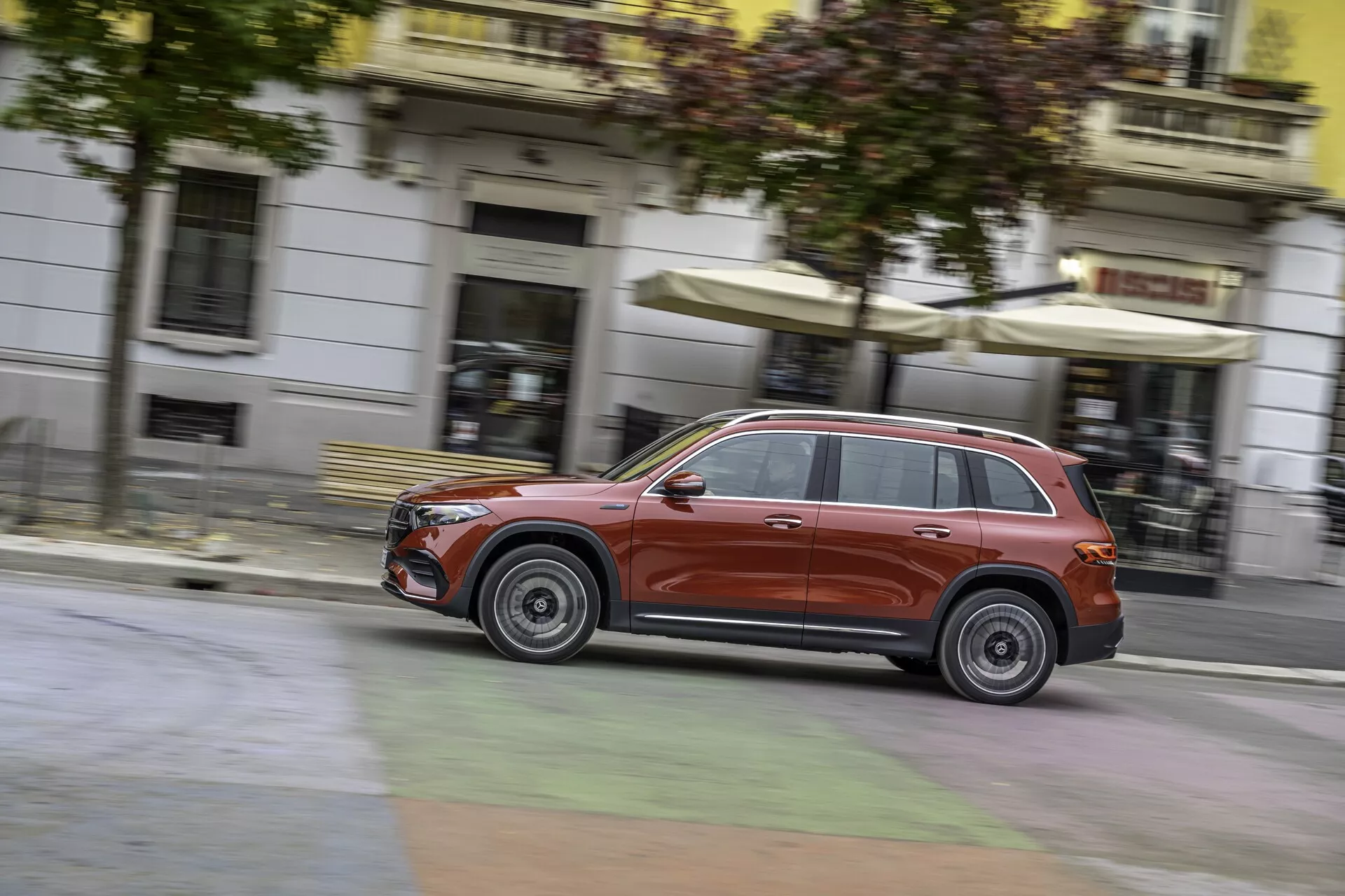 Dynamisk sidovy av en metallic röd Mercedes EQB 2021 SUV i rörelse, som framhäver dess distinkta designelement som frontgrill, strålkastare och lättmetallfälgar. Rörelseoskärpan i bakgrunden och på hjulen betonar dess hastighet och smidighet genom en stadsmiljö.