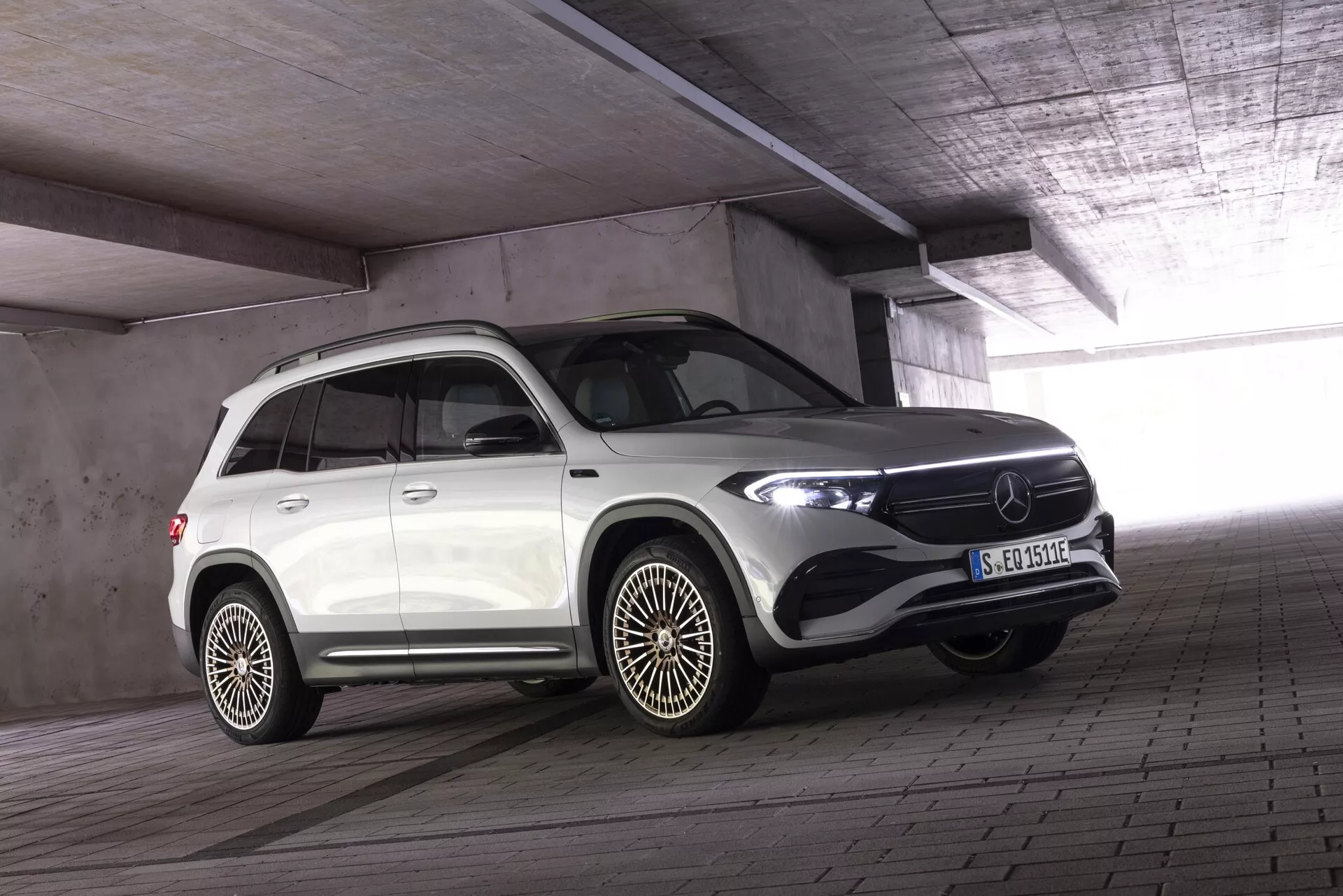Vit lyxig Mercedes EQB 2021 SUV i inomhusparkering med betongtak och pelare, fram och sida trekvartsvy; LED-strålkastare, framträdande grill med stor emblem och el-/hybridskylt på skärmen. Fälgar av legering med flerspokesdesign och skyddskant på dörrarna som indikerar off-road-design. Fordonet är parkerat med framhjulen något vända mot kameran, vilket ger en dynamisk komposition.
