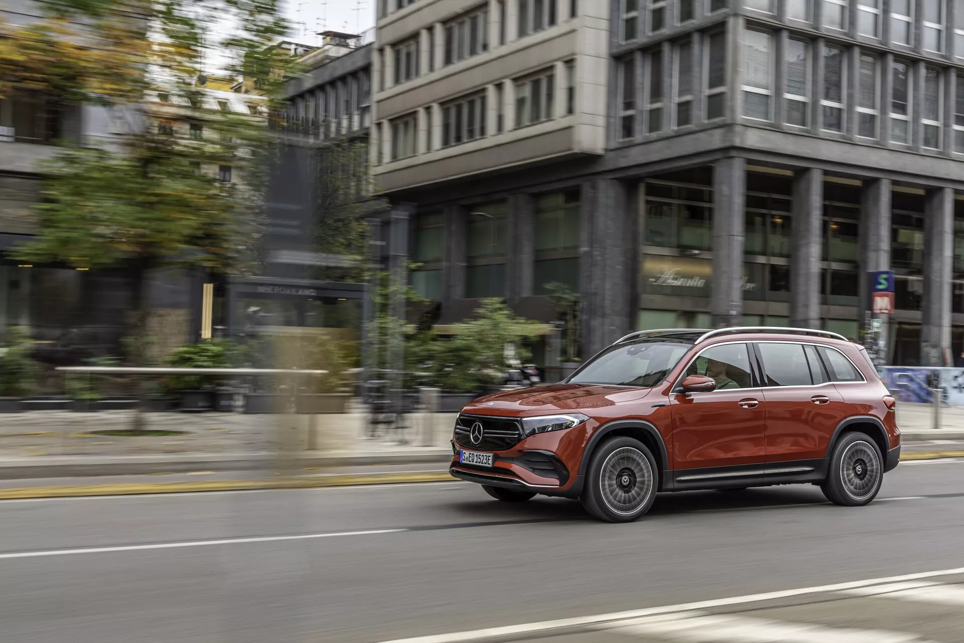 Bildens motiv är en röd Mercedes EQB 2021 SUV i rörelse, fångad från höger sida i en dynamisk panoramabild. SUV:n kör på en stadsgata med byggnader och trottoar i bakgrunden och har ett glansigt yttre, stiliga lättmetallfälgar, krominlägg runt fönstren, takräcken och integrerade blinkers i sidospeglarna. Den något framåtvinklade vyn framhäver den främre passagerarsidan och visar avancerade strålkastare mot en suddig stadsbakgrund, vilket skapar en känsla av fart och elegans.