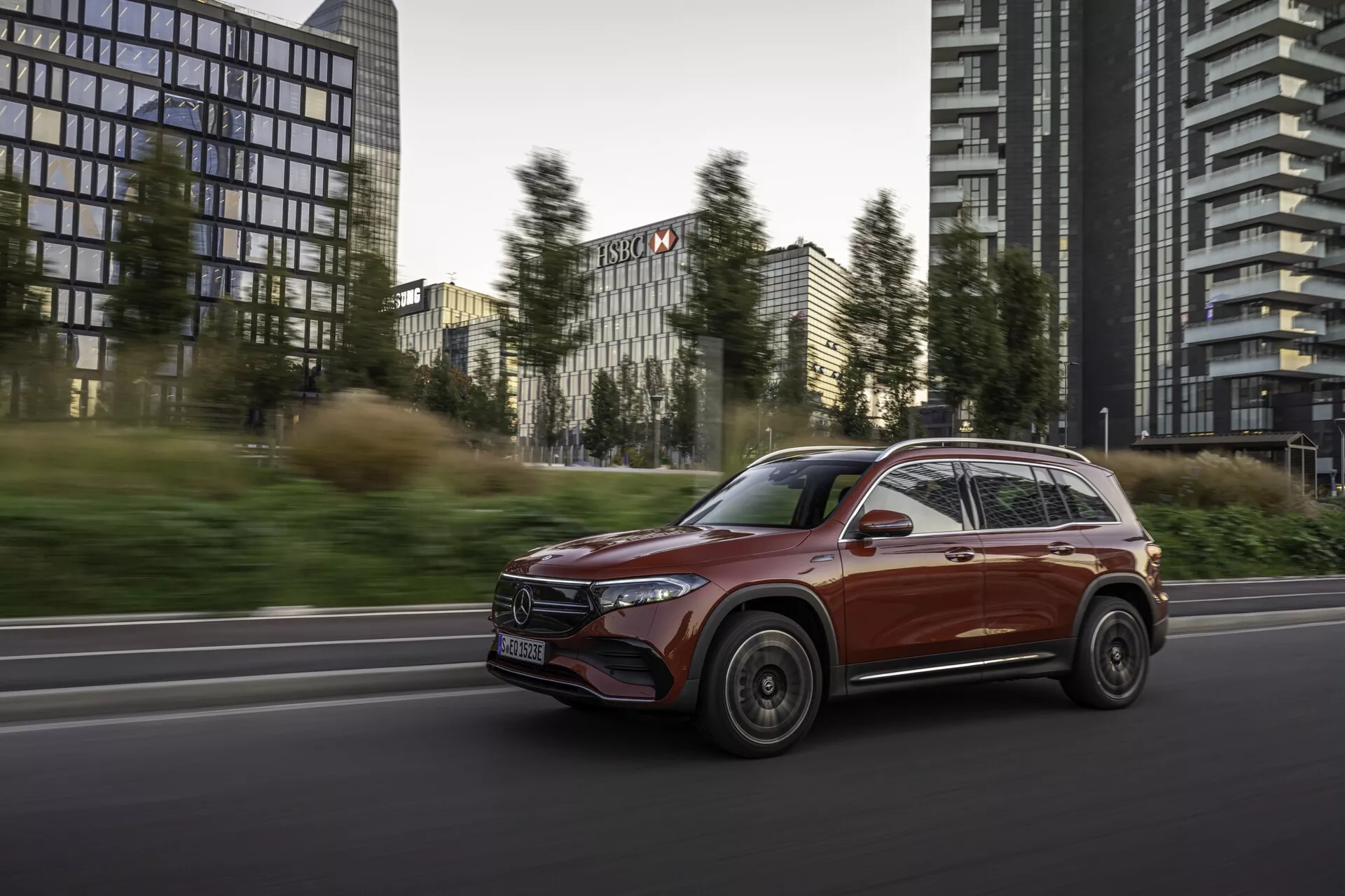 Dynamisk sidovy framifrån av en 2021 Mercedes EQB SUV i rörelse på en stadsväg med kontorsbyggnader i bakgrunden. Fordonets eleganta, aerodynamiska design, glänsande finish och avancerade funktioner såsom moderna LED-strålkastare och integrerade körriktningsvisare på sidospeglarna framhävs. Mjuk belysning accentuerar bilens kombination av lyx och funktionalitet med rörelseoskärpa på hjulen och bakgrunden som tydligt fastställer dess hastighet och riktning.