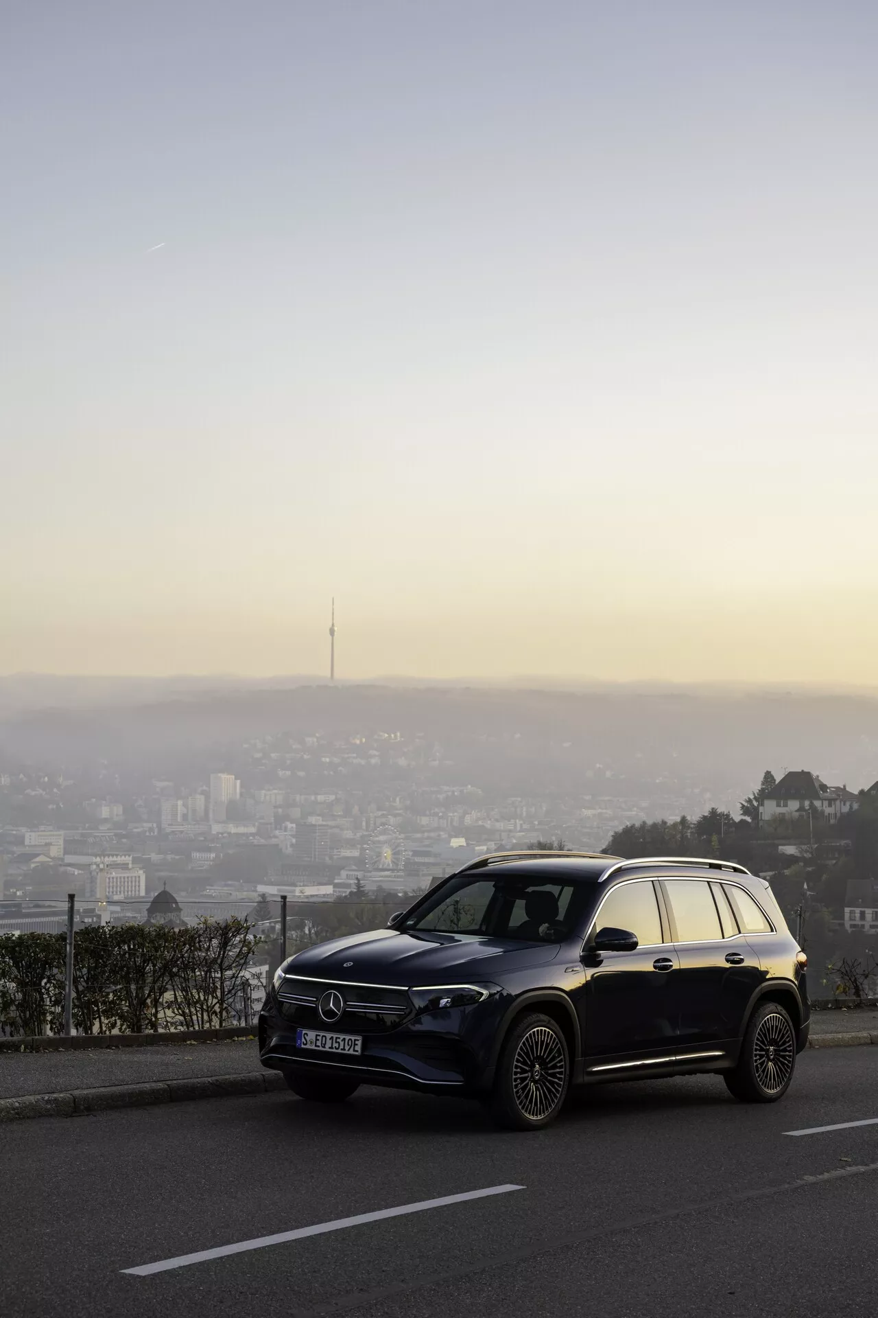 En modern Mercedes EQB 2021 SUV placerad på en utsiktspunkt med utsikt över en stad i skymningen, fångad i en trekvartsvinkel framifrån som visar upp dess LED-strålkastare, framträdande främre grill med stort emblem, lättmetallfälgar och en blank metallisk eller pärlemorfärg. Fordonet är skarpt mot en suddig stadsbakgrund, fångat under den gyllene timmen med en himmel som övergår från blått till varma nyanser.