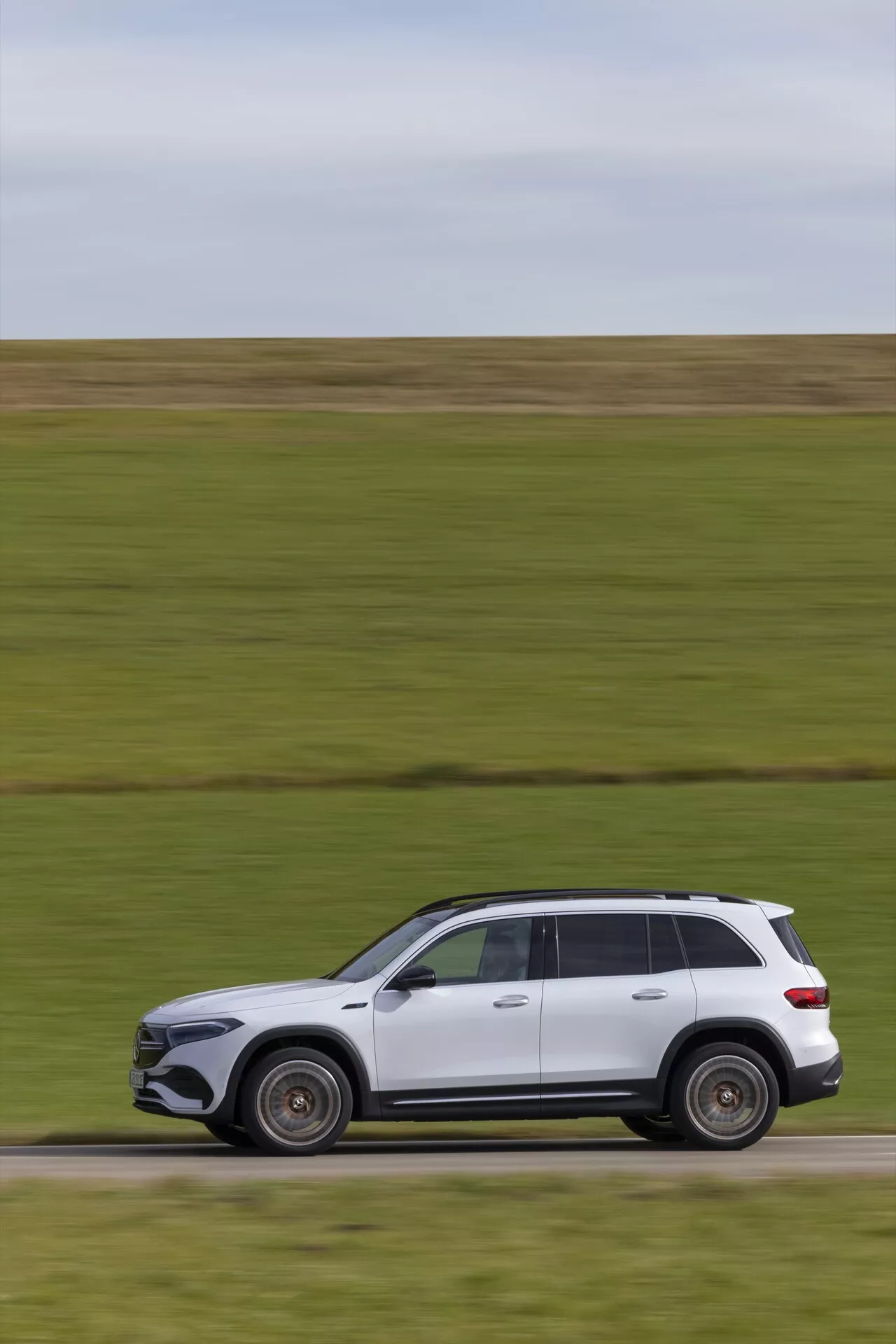 Sidoprofil av en modern lyx-SUV i rörelse, sannolikt en nyare modell Mercedes EQB 2021, målad i vitt. Fordonet, som rör sig från vänster till höger, visar sofistikerade designelement med en elegant aerodynamisk kaross, lättmetallfälgar, kromlister runt fönstren och takräcken. Trots den suddiga bakgrunden som antyder hastighet, förblir bilen i skarpt fokus och framhäver dess felfria lack och dynamiska hållning.