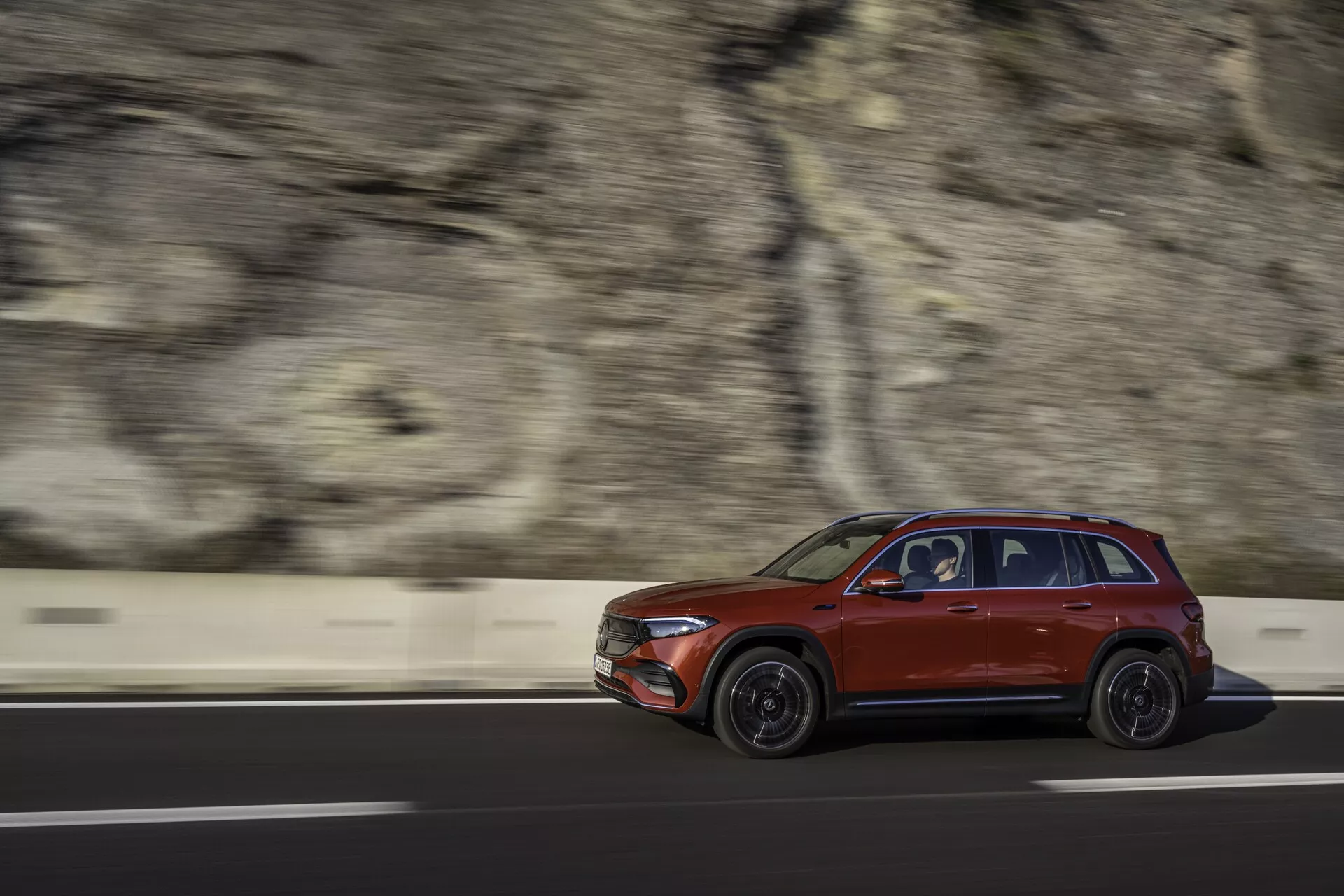 Dynamisk sidoprofilbild av en röd 2021 Mercedes EQB SUV i rörelse, kör från vänster till höger på en väg med en suddig bakgrund som visar en barriär och en klippvägg. Fångad med snabb slutartid, är fordonet skarpt fokuserat och visar designfunktioner som dörrarnas former, hjuldesignen, sidospeglarna, fönstren och ett takräcke under mjukt morgon- eller eftermiddagsljus.
