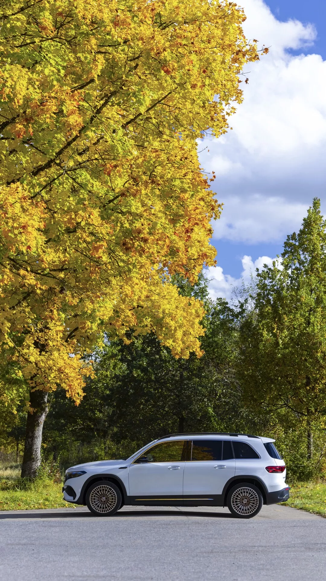 Sidoprofilvy av en lyxig Mercedes EQB 2021 SUV parkerad parallellt på en tvåfilig väg, med ett tvåfärgat färgschema med ljus kropp och mörkare nedre trim, högklassiga flerspoksfälgar, terrängdäck och omgivet av en lugn naturlig miljö med träd som har gult lövverk, vilket antyder höst.