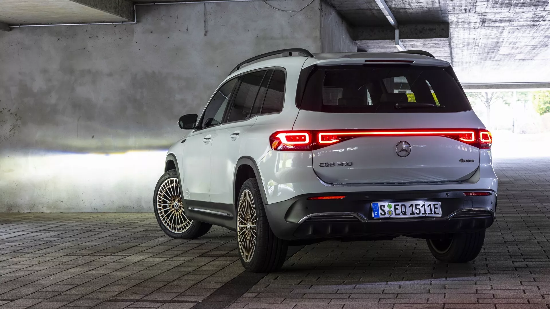 Trebaksvy från sidan av en vit Mercedes EQB 2021 parkerad i en inomhusmiljö, troligen ett garage. Fordonets bakre design, inklusive baklucka, baklampor och emblem med en elektrisk symbol, är fullt synliga. Passagerarens sidoprofil visar bakdörren och en del av framdörren, aerodynamiskt optimerade hjul med intrikata ekerdesigner och en europeisk stadsnummerplåt. Bakre lyktor lyser upp och framhäver ljusdesignen. Den vita glansiga finishen kontrasterar mot betongstrukturen, konstgjord belysning och målade golvmarkeringar i inomhusmiljön.