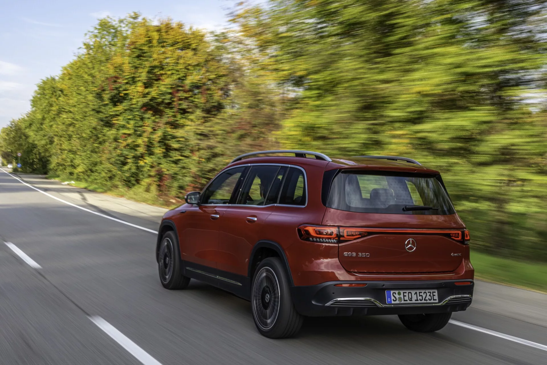 En modern SUV Mercedes EQB 2021 (243) i metallic röd, fångad från en bakre trekvarts vinkel på en väl underhållen förorts- eller landsväg. Bilens eleganta och sportiga design lyfts fram av en sluttande taklinje, kort bakre överhäng, och en karaktärslinje längs sidan. Synliga funktioner inkluderar stora flerkantiga lättmetallfälgar med lågprofildäck, strömlinjeformade LED-bakljus och bakre parkeringssensorer. Bilden förmedlar en känsla av dynamisk körning med fordonet i rörelse och bakgrunden något suddig med trädridåer.