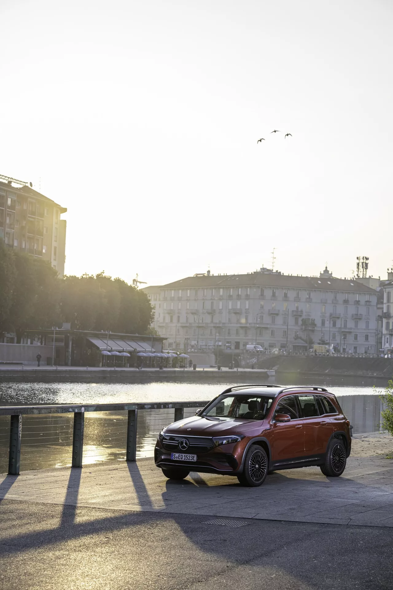 Bilden visar en elegant Mercedes EQB 243 SUV från 2021 parkerad vid kanten av ett stadsvattenområde. Fordonet fångas från en delvis framifrån och sidovinkel, vilket framhäver vänstra sidan och vänster främre kvadrant mot en lugn vattenscen med avlägsna byggnader. Nyckeldesignelement som strålkastarnas form, den främre stötfångaren med integrerade dimljus eller sensorer, grillens mönster och motorhuvens linjer är tydligt synliga. Scenen badar i varmt solljus och framhäver fordonets övre designfunktioner och taklinje. Vindrutan och de främre fönstren ger insyn i den halvgenomskinliga kabinen. Bilens avancerade belysningssystem, sensorer i den nedre delen av främre stötfångaren, hög markhöjd, lättmetallfälgar och en eventuell panoramatak antyder lyx- och avancerade tekniska funktioner. Metalliclackeringen speglar fordonets nya eller väl underhållna skick.