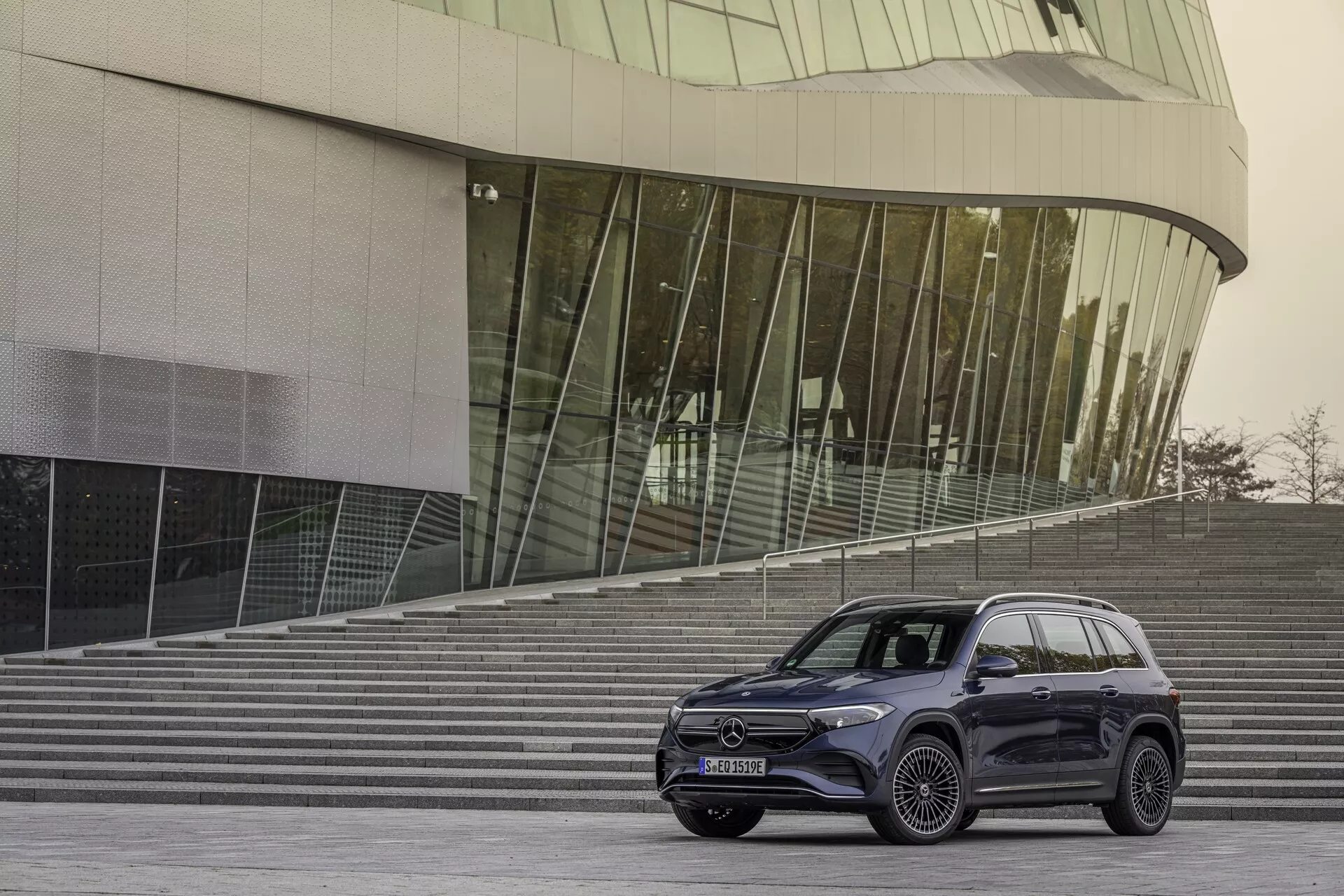 Trekvarts främre vy av en Mercedes EQB 2021 (243), som visar sidoprofilen och fronten. Fordonet, placerat 10-15 fot från kameran, är fotografiets mittpunkt. Det står på ett stenlagt område med arkitektoniska trappor och en modern glasklädd byggnad i bakgrunden, vilket ger en urban och samtida känsla. Fronten inkluderar eleganta LED-strålkastare, en framträdande grill med ett stort emblem och en del av stötfångaren. Sidan visar flerspokes fälgar med stor diameter och låga däckprofiler, vilket indikerar en lyxig eller sportig trim. SUV har släta karosslinjer och en mörk, blank yta, möjligen i grafit eller svart. Fönstren är tonade och sidospeglarna har integrerade blinkers. Taklinjen lutar något och har praktiska takräcken. Troligen inkluderar den avancerade teknologiska funktioner som adaptiva strålkastare och parkeringssensorer, även om dessa inte är tydligt urskiljbara.