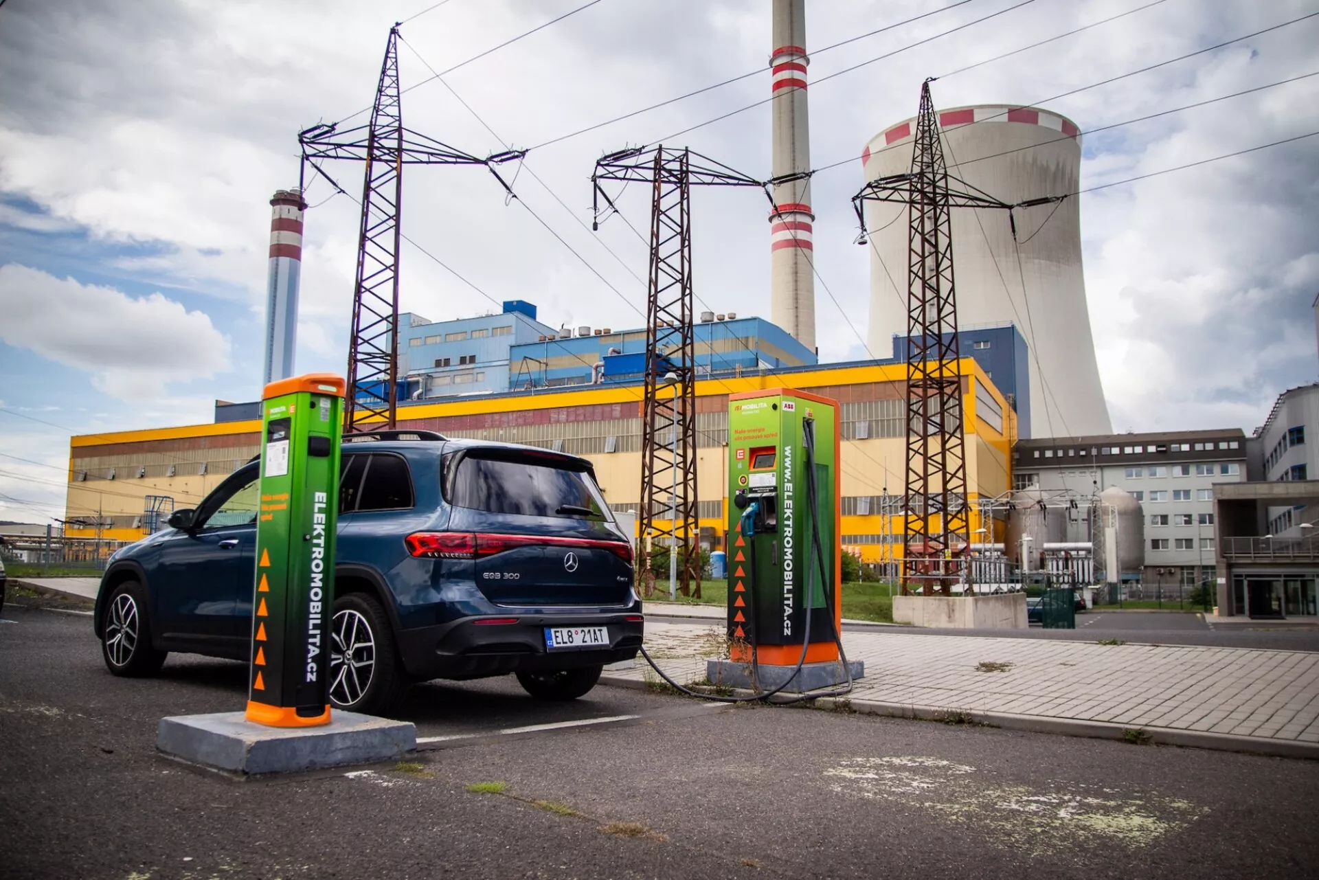 Elektrisk hatchback Mercedes-Benz EQB 2021 parkerad vid en laddningsstation med en inkopplad kabel, sedd från ett tre kvarts bakperspektiv, som framhäver dess bakljus, fönster och passagerarsida mot en industriell bakgrund