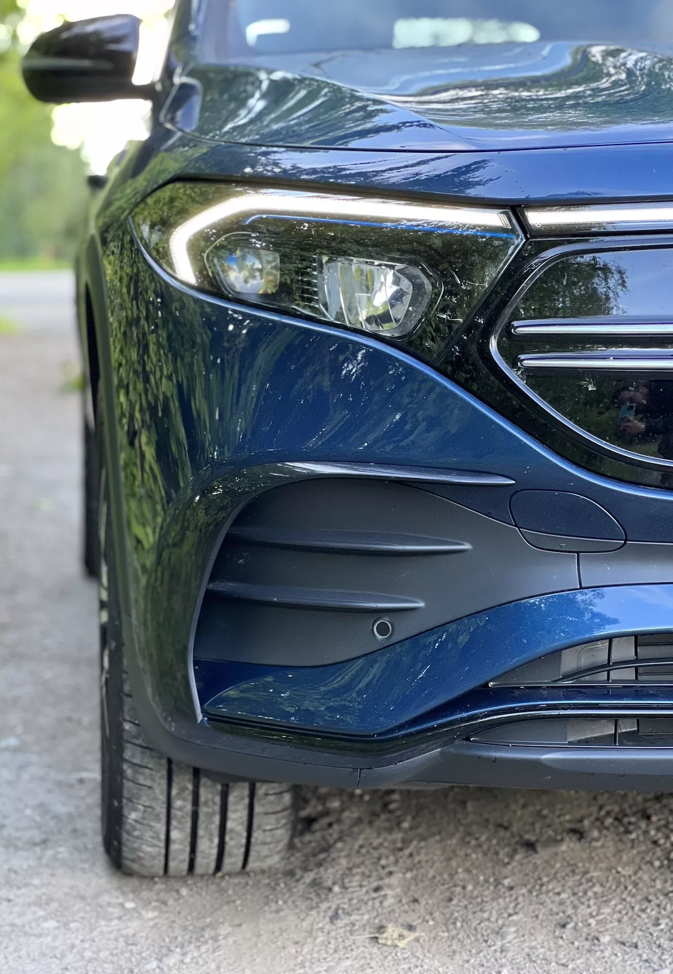 Närbild av främre vänstra sidan av Mercedes-Benz EQB 2021, med LED-strålkastare, del av grillen med plats för emblem, aerodynamiskt utformad frontstötfångare med luftintag, flerspakigt lättmetallhjul med detaljerat mönster på däcket, och metallisk målning som reflekterar sofistikerade karosskurvor