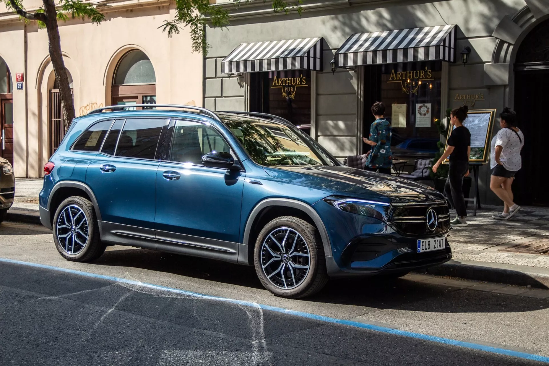 Sidoprofil av Mercedes-Benz EQB 2021 på stadsgata, som visar modern lyxig SUV-design med stora aluminiumfälgar, integrerade blinkers i sidospeglarna, tonade rutor som reflekterar den urbana miljön, släta karosslinjer, takräcken och dörrhandtag i linje med karossen i naturligt ljus