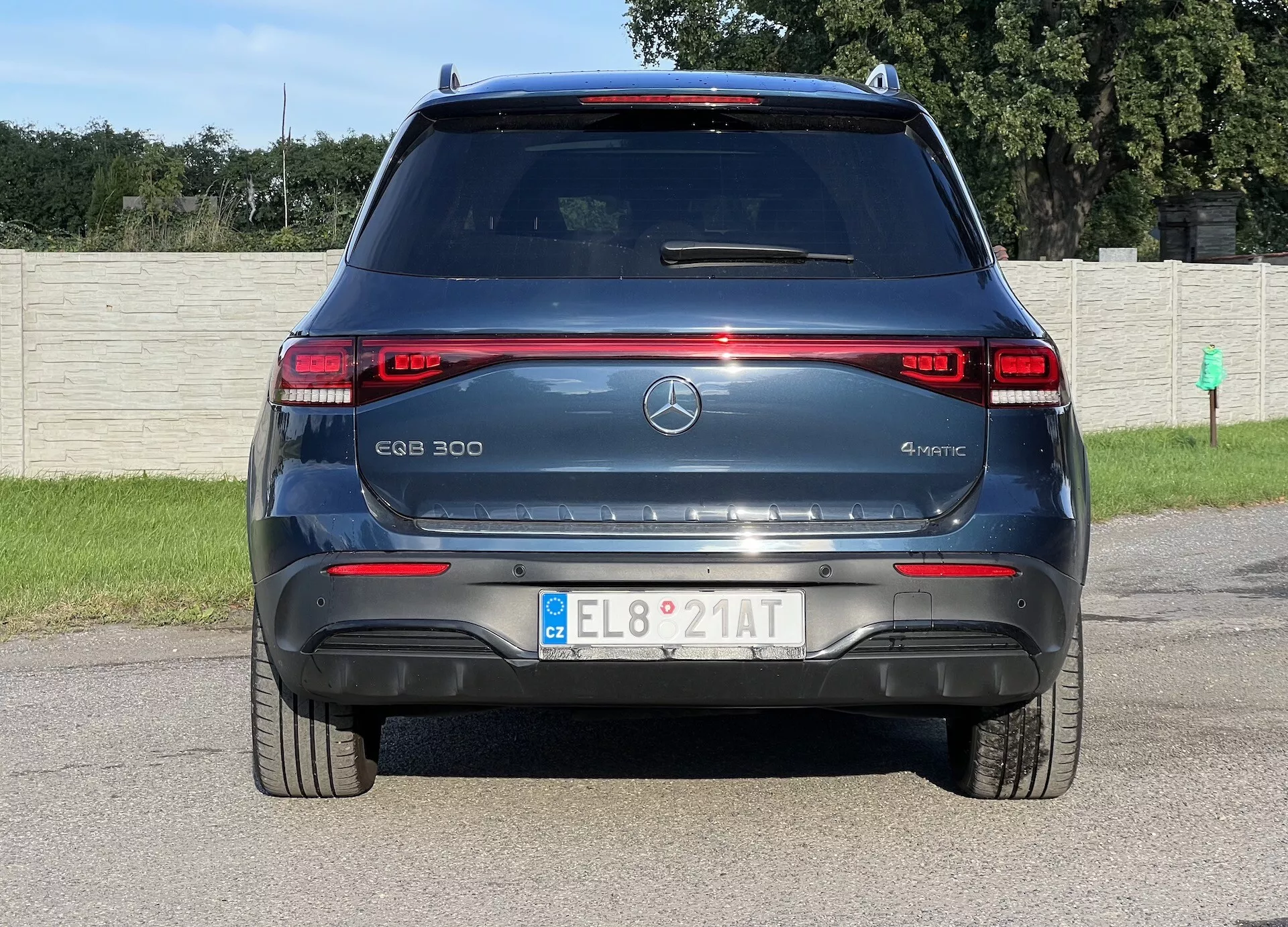 Bakvy av el-SUV:en Mercedes-Benz EQB 300 4MATIC som visar LED-bakljus, model och elektrisk märkning, trestjärnig emblem, bakruta med avfrostare och torkare, parkeringssensorer, röda reflexer, europeiskt registreringsskylt, aerodynamisk bakspoiler och 4MATIC fyrhjulsdriftsystem, mot en bakgrund av ett gräsfält och staket.