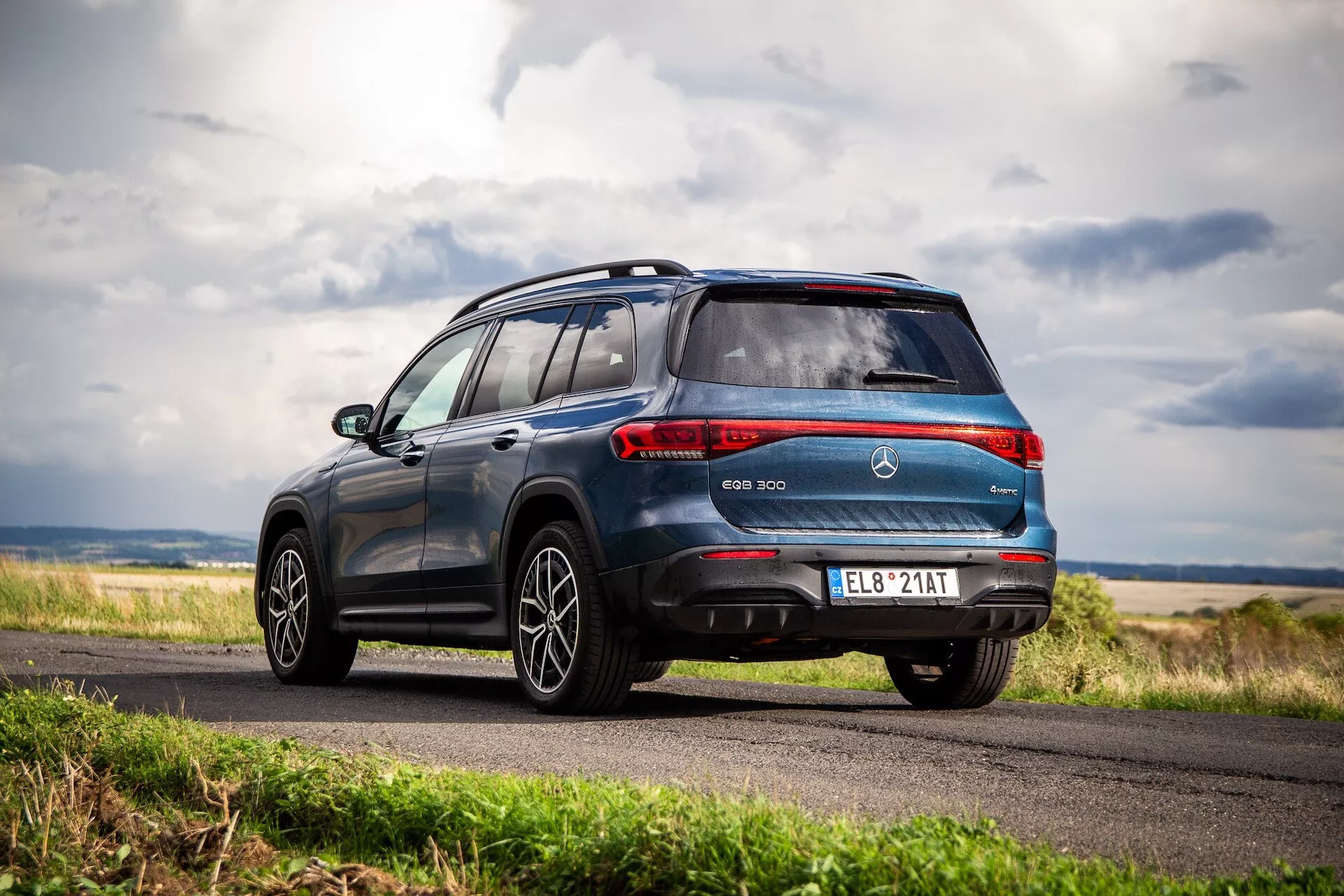 2021 Mercedes-Benz EQB elektrisk SUV visas från en dynamisk bakre tredjedels vinkel som visar fordonets slanka design, inklusive bakre kombinationslampor, en sportig takspoiler, legeringshjul och EQB-märkning mot en bakgrund av en molnig himmel och lantligt landskap.