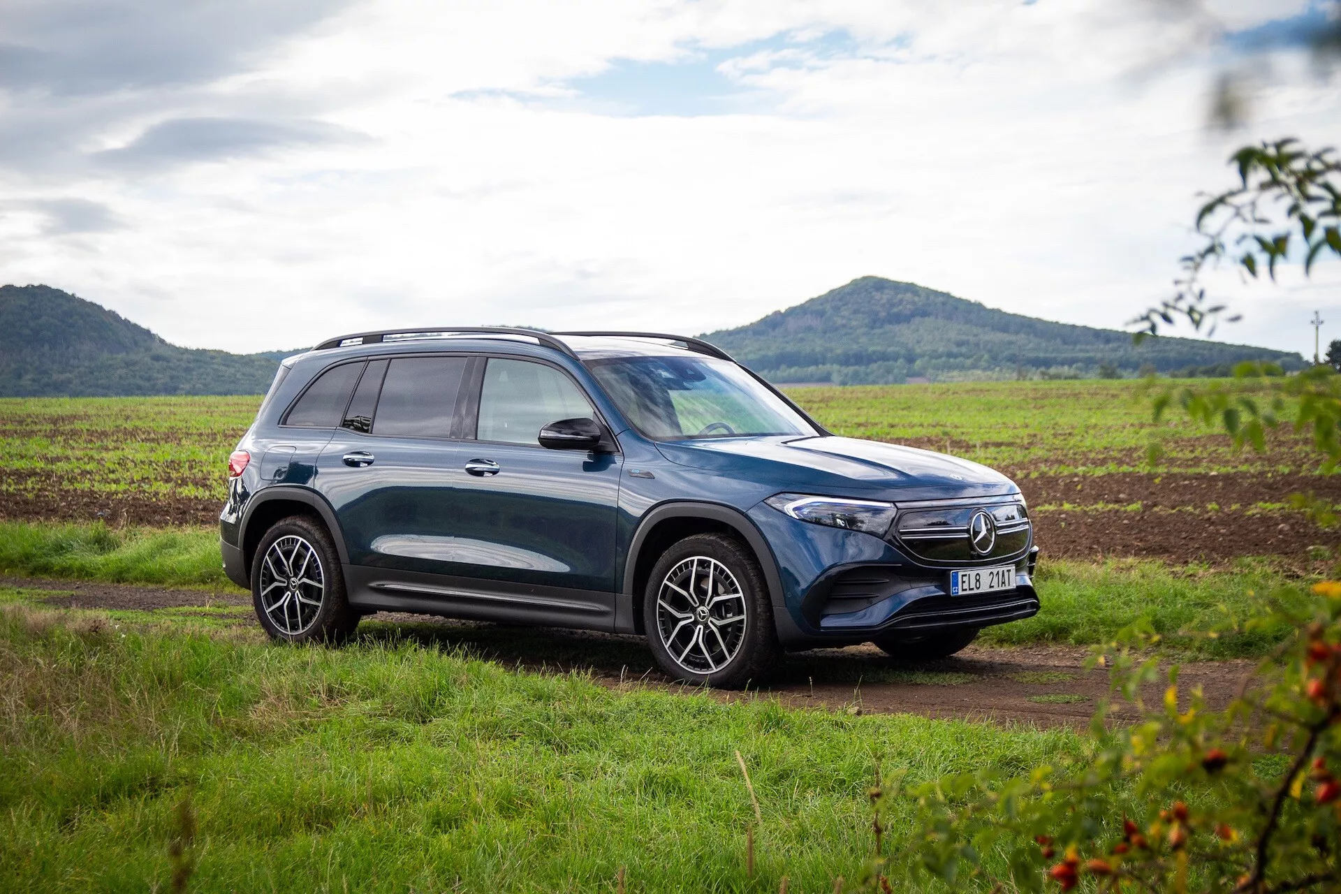Sidoprofil av en mörkfärgad SUV Mercedes-Benz EQB 2021 parkerad på gräsmark med en grusväg framför, visar upp legeringshjul, takräcken och sportiga designdrag mot bakgrund av ett lantligt landskap med delvis molnig himmel