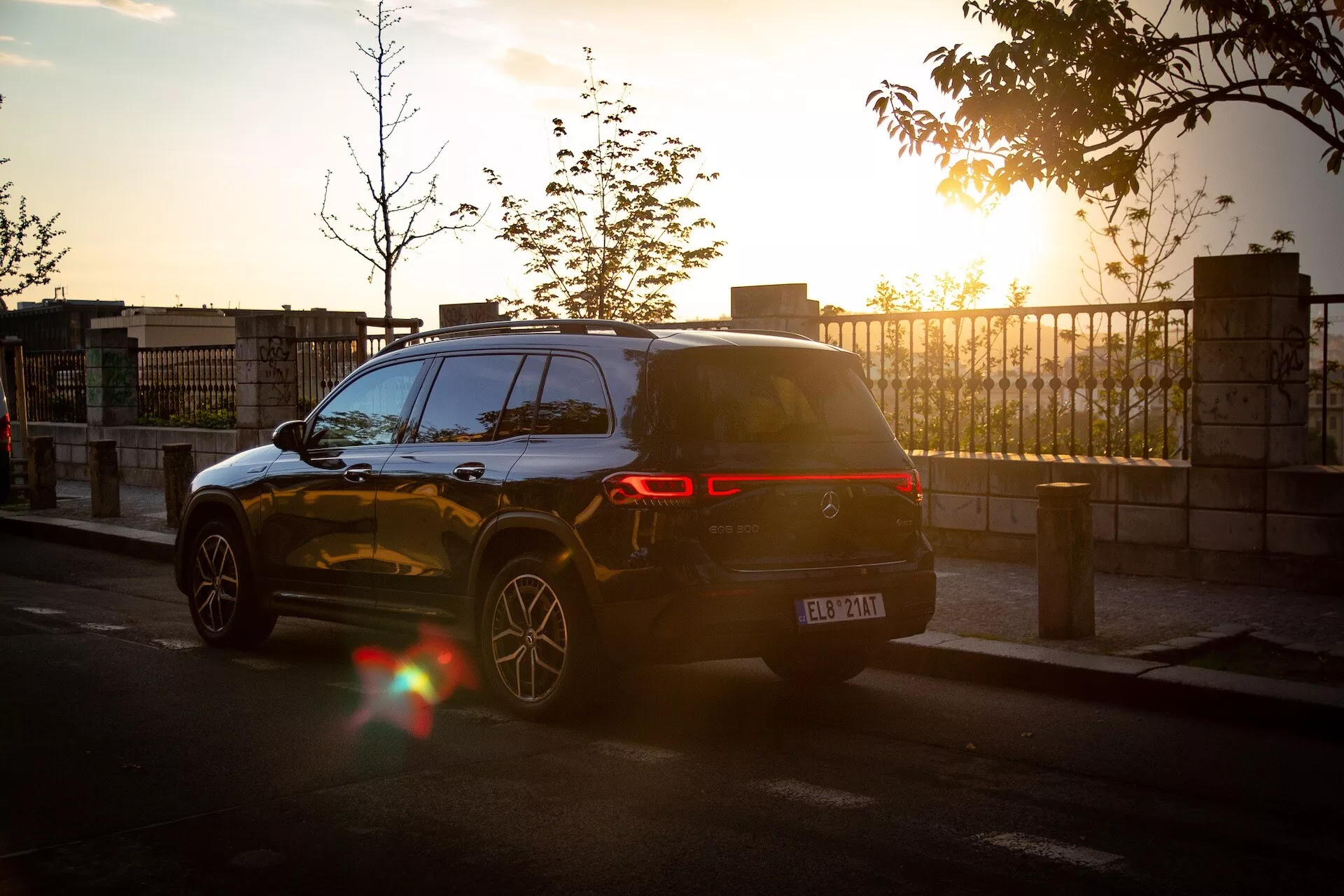 Mercedes-Benz EQB 2021 i en dynamisk bakre trekvarts vy vid solnedgången med objektivglans, som visar dess moderna SUV-design, LED-baklyktor, parkerad på en stadsgata som reflekterar det varma solljuset från dess blanka, mörkfärgade lack och flerspakiga legeringshjul, inklusive parkeringssensorer synliga på bakre stötfångaren