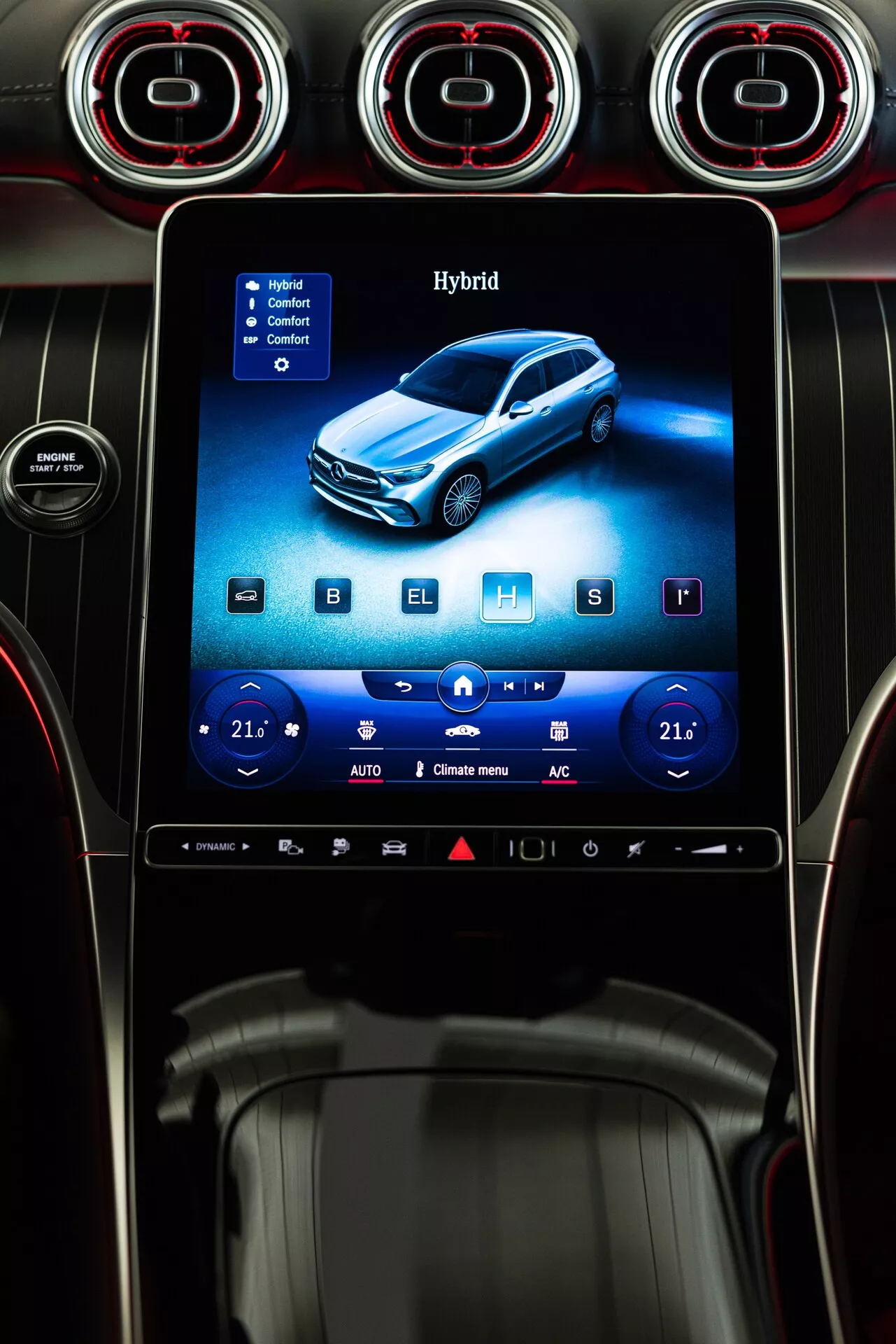 Närbild av den centrala konsolen i en 2022 Mercedes GLC 254, med ett upprättstående infotainmentsystem med pekskärm inramat av tre cirkulära luftventiler med röda accentringar. Skärmen visar ett blått hybridfordon, med digitala klimatkontroller inställda på 21 grader Celsius på båda sidor, vilket indikerar en tvåzons klimatanläggning. Ikoner för stolsvärme, kylning och avfrostningsalternativ är synliga, tillsammans med en snabbåtkomstlist längst ner. Konsolen är designad med borstad metall, glansiga svarta ytor och minimalistiska knappar, vilket framhäver en modern och lyxig interiör.
