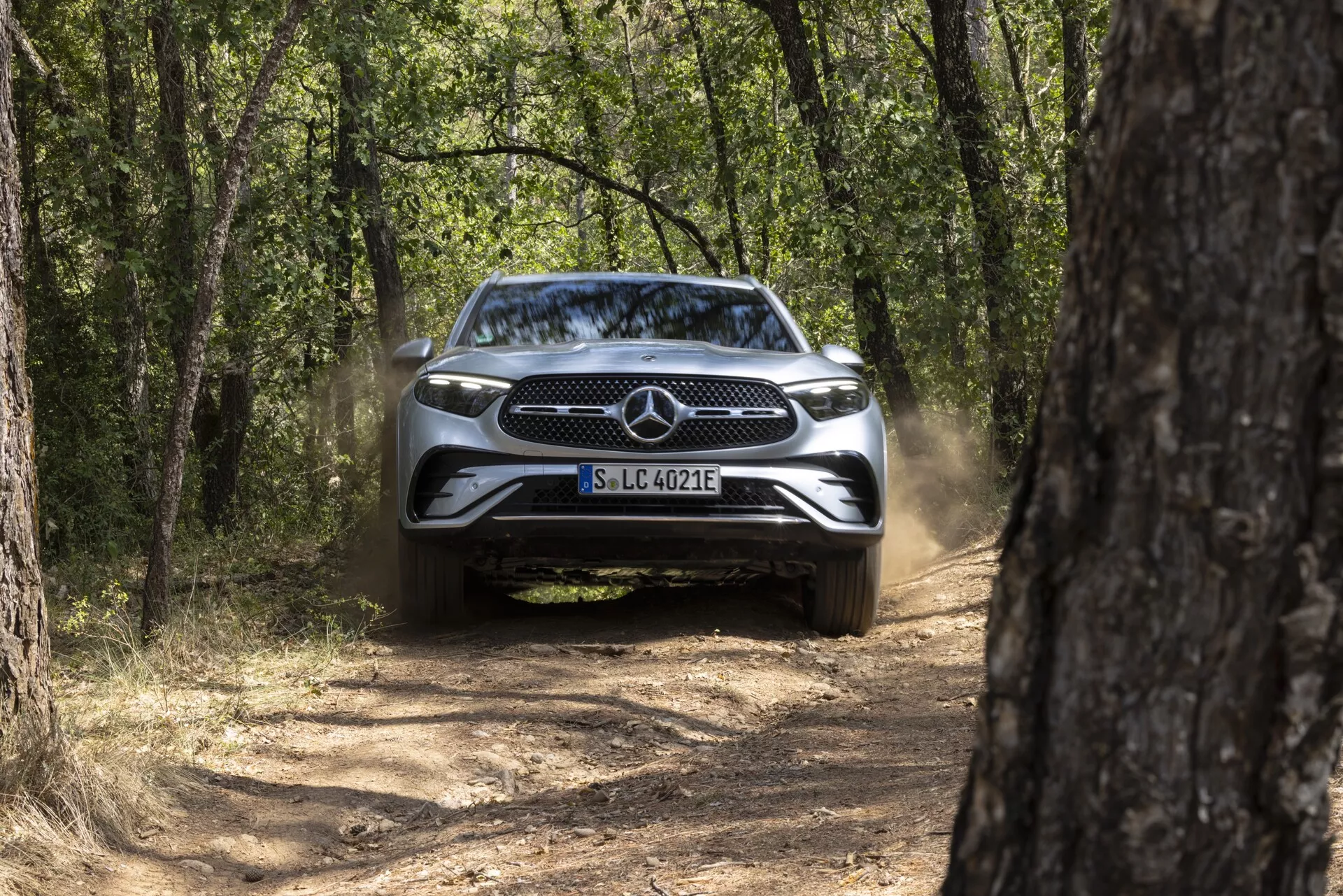 Främre vy av en Mercedes GLC 2022 (254) på en smal grusväg, omgiven av träd, utan tända strålkastare, damm bakom, solig dag med naturligt ljus.