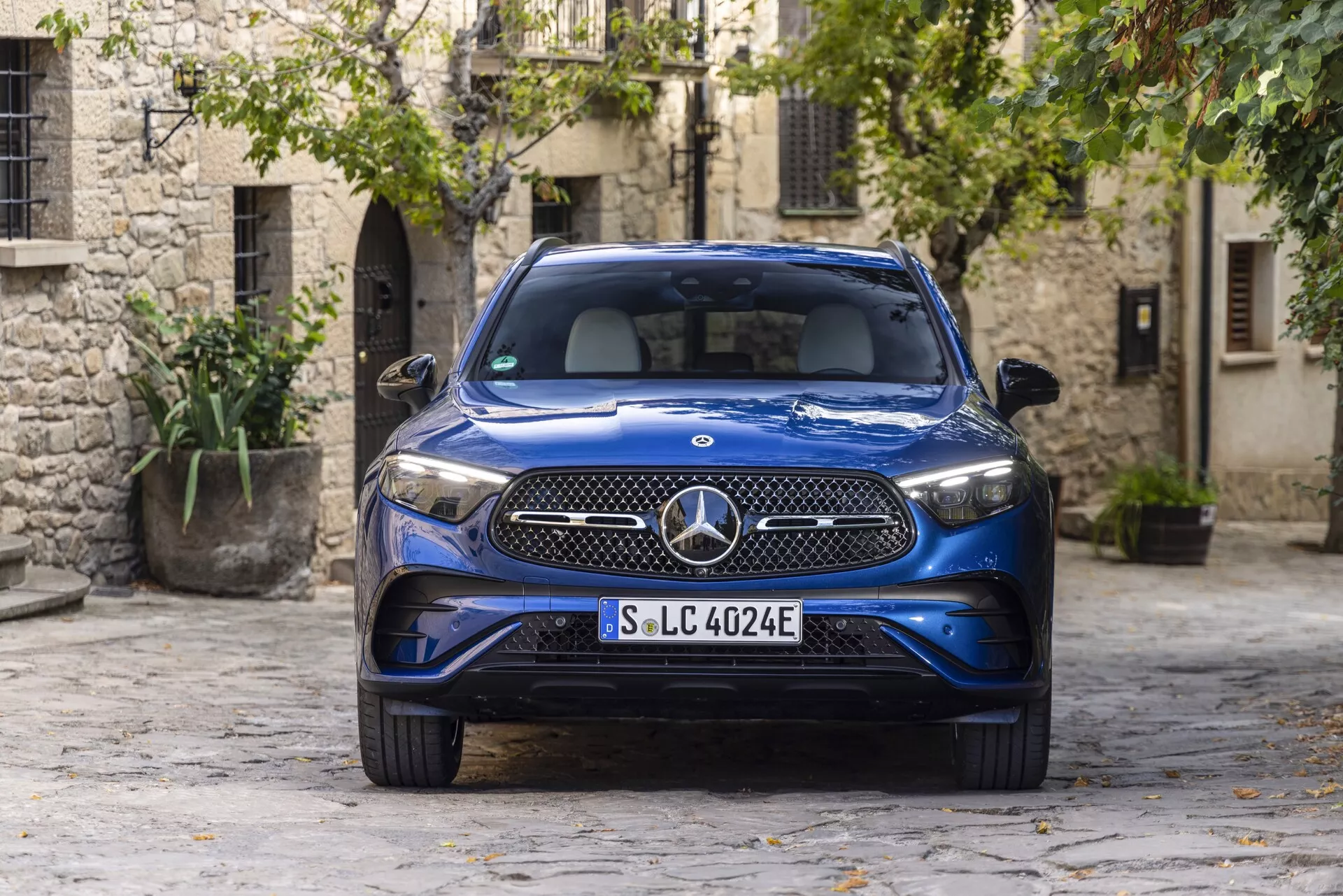 2022 Blå Mercedes GLC 254 lyx-SUV parkerad i en pittoresk kullerstensbelagd innergård inom en gammal stadsdel, visar upp LED-strålkastare, distinkt frontgrill med stor Mercedes-Benz-emblem, EQ-märke som indikerar elektrisk eller plug-in hybrid natur, elegant metallisk exteriör och integrerade luftventiler i den främre stötfångaren. Fotograferad från en frontvinkel, som framhäver modern biltillverkning mot traditionella stenbyggnader och grönska.