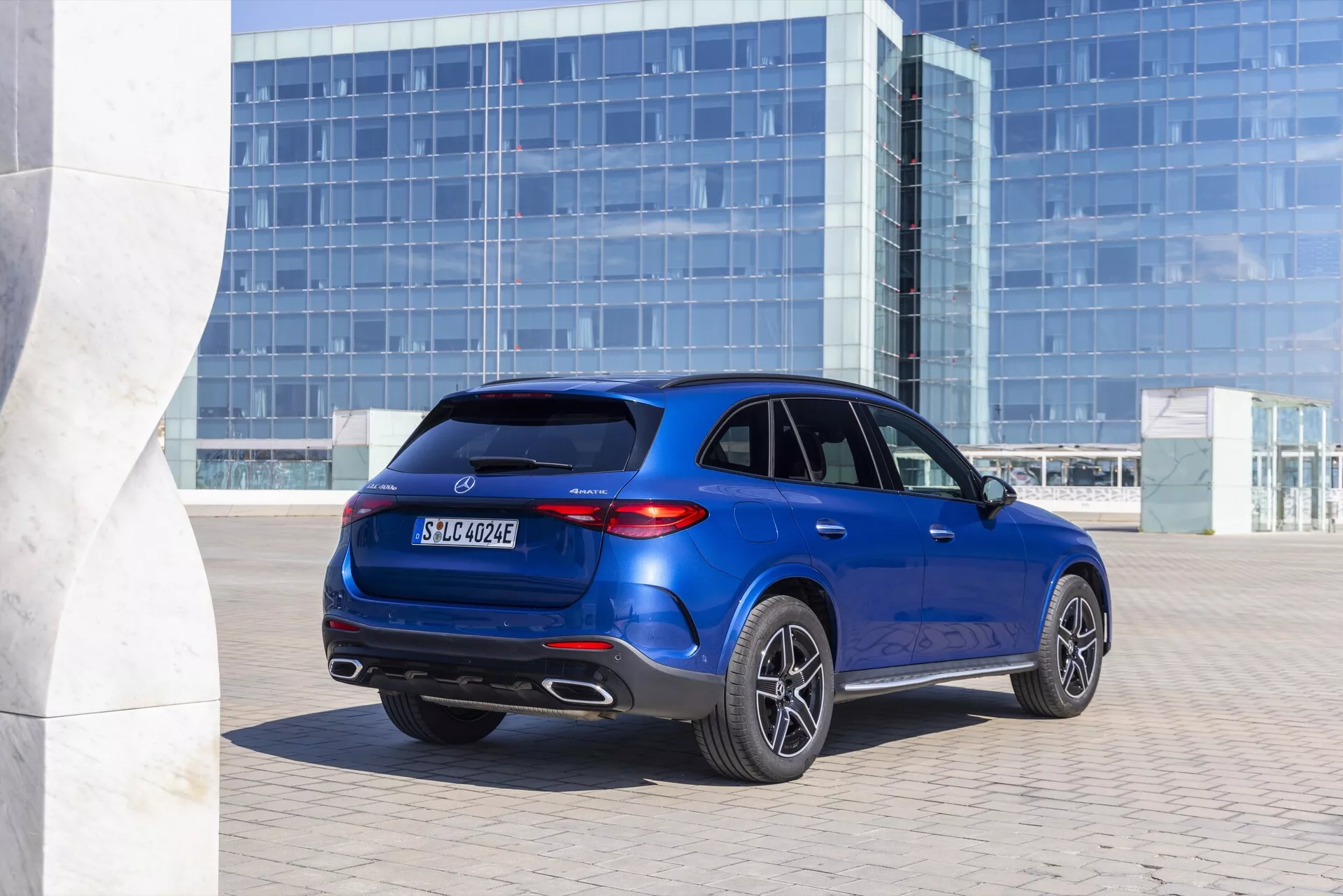 Trekvart bakre vy av en blå Mercedes GLC 2022 SUV som visar sidans och bakre design, bakljus, dubbla avgasrör, reflexer på bakre stötfångaren, takspoiler och lättmetallfälgar. Fordonet är parkerat framför en modern glasfasad, vilket tyder på en urban miljö. Bilens bakre emblem indikerar att det kan vara en elektrifierad modell, sannolikt hybrid eller helt elektrisk.