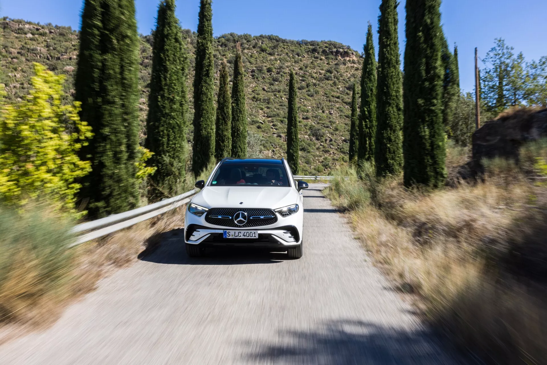 Dynamisk bild av en bil från frontperspektiv, en Mercedes GLC 2022 (254), som kör mot kameran på en solbelyst, smal väg omgiven av höga träd. Visar bilens hela front, inklusive grill med Mercedes-Benz-emblem, eleganta LED-strålkastare, motorhuv, vindruta och del av taket. Den lätta oskärpan på hjulen och det streckade bakgrunden skapat av rörelseoskärpa-teknik framhäver bilens rörelse och dess avancerade funktioner, såsom möjlig LED-strålkastarteknik, uppdaterad grilldesign med aktiva aerodynamiska element eller sensorer för förarassistanssystem och troligtvis parkeringssensorer eller kameror.